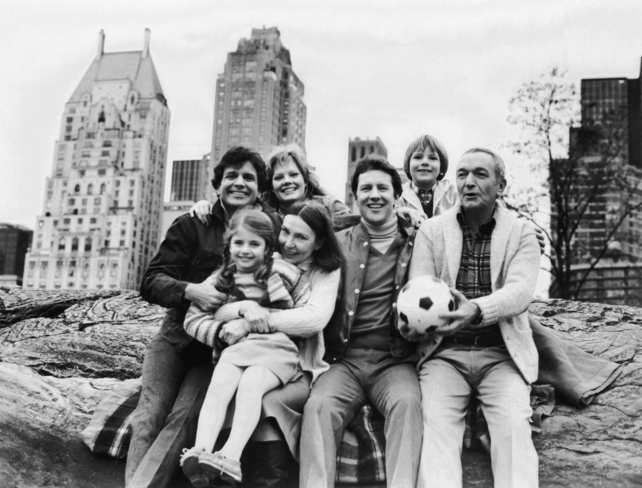 A group of adults and two children smile in a black and white image with a cityscape in the background.