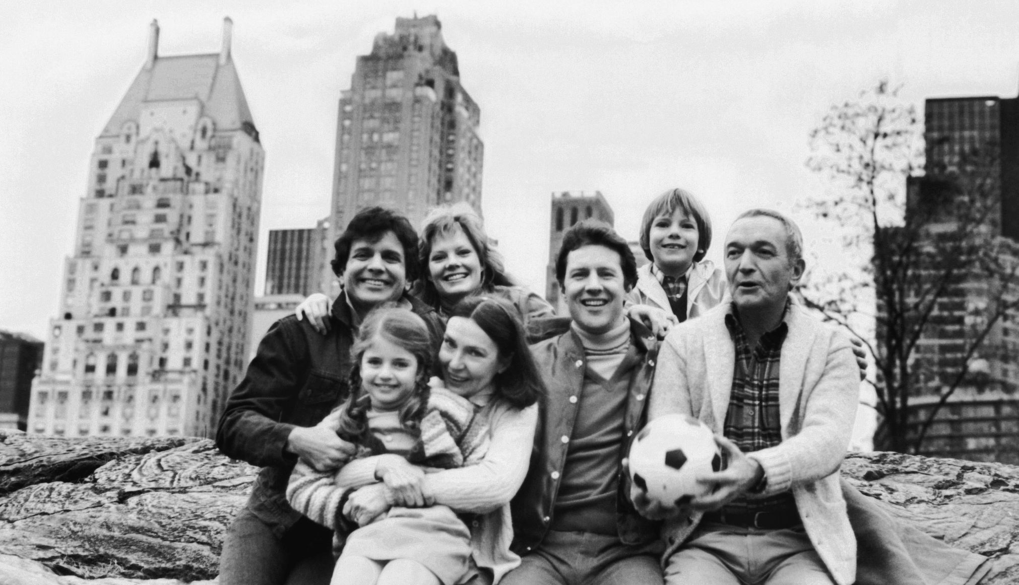 A group of adults and two children smile in a black and white image with a cityscape in the background.