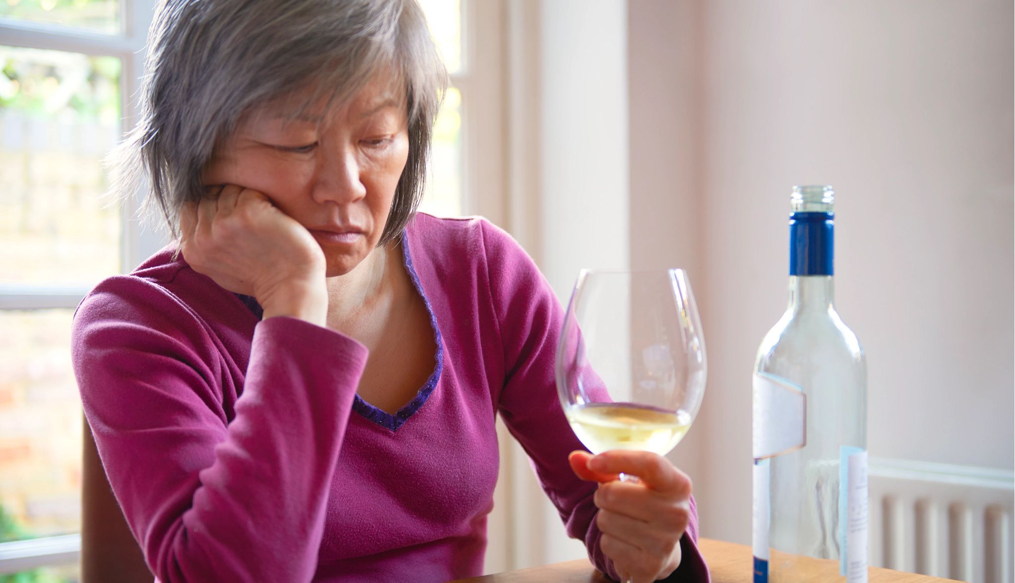 Drinking alcohol during menopause is an unhealthy combination for mood and memory A woman sitting at a table with one hand on her chin and the other holding a glass of wine