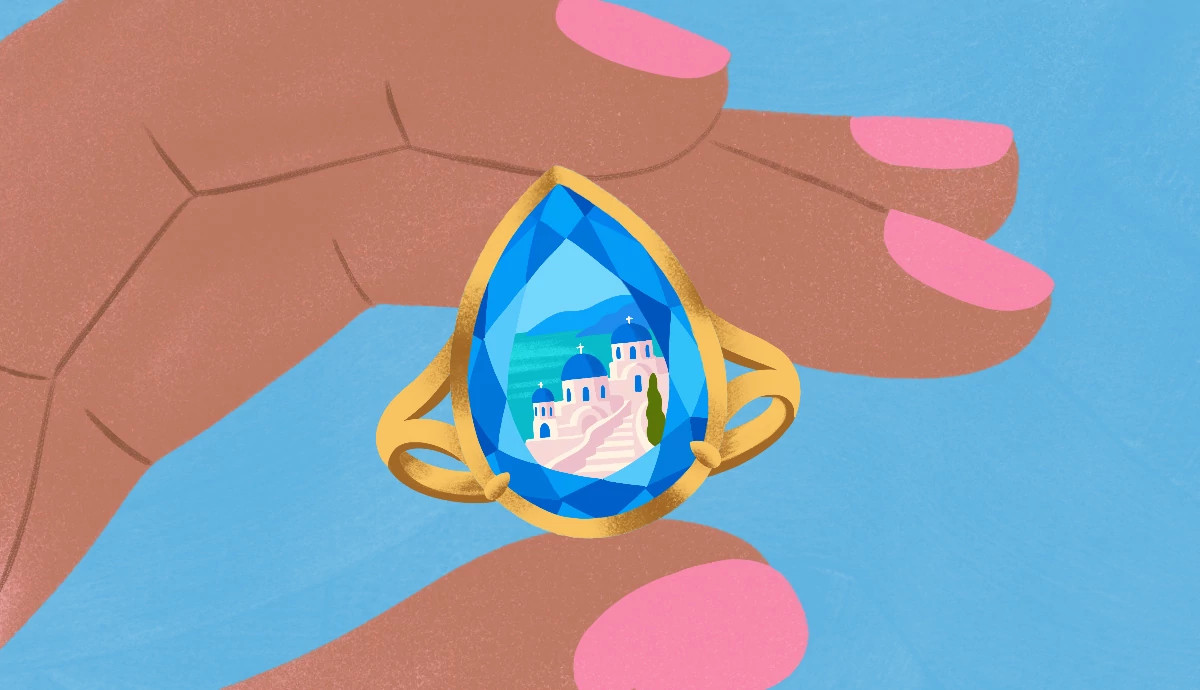 Hand holding a gold ring with a tear-shaped blue gemstone reflecting white houses with blue domes on a coastal hill.