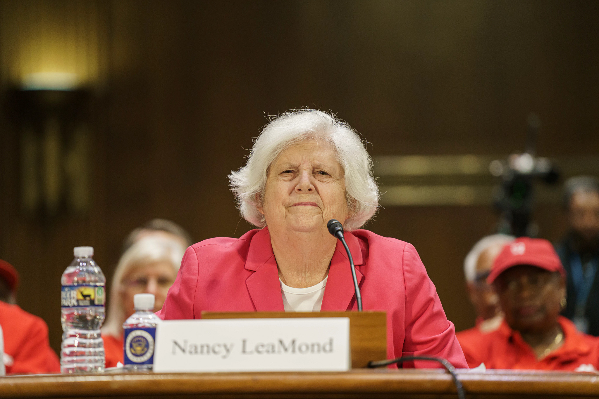 A woman in glasses, a red blazer and white shirt, sits in front of a microphone.