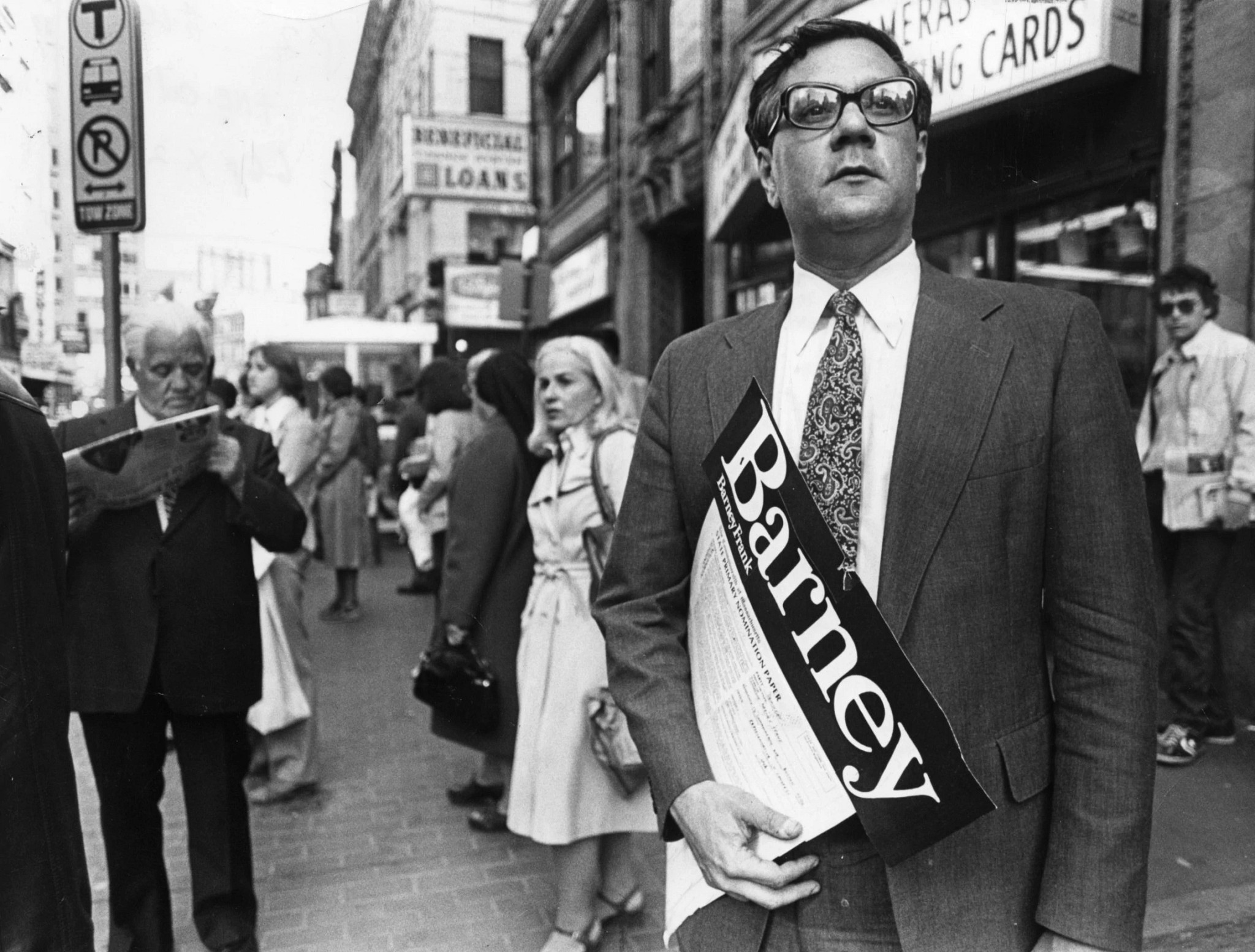 barney frank standing on a street with a poster