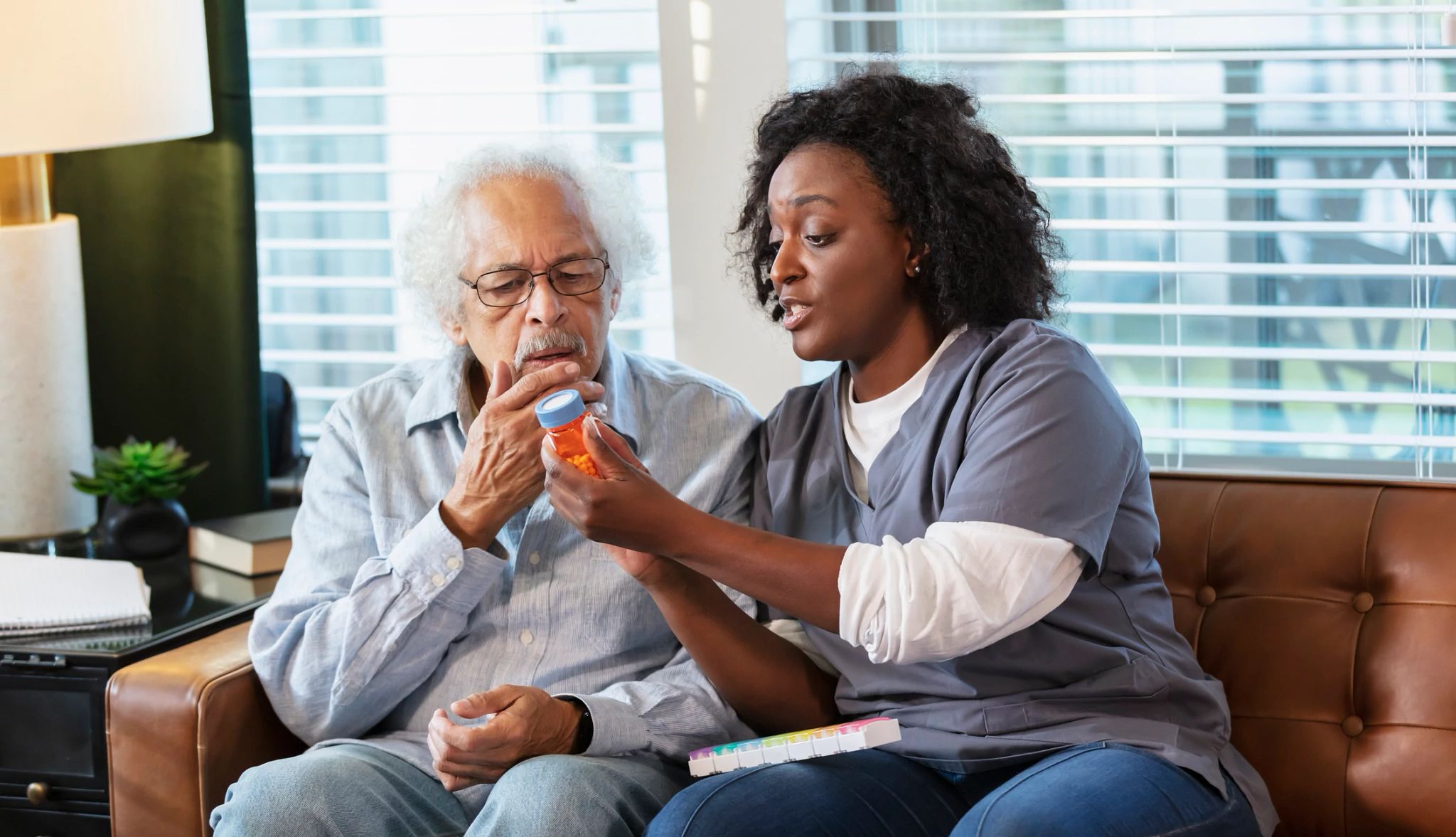 Un asistente de atención médica a domicilio ayuda a una adulto mayor con su medicamento y le explica cuándo debe tomarlo. La trabajadora de la salud es una mujer afroamericana de unos 30 años. Su paciente es un hombre multirracial hispano y caucásico. Están en su casa, sentados en su sofá, ambos mirando el frasco de pastillas. Un asistente de atención médica a domicilio ayuda a una adulto mayor con su medicamento y le explica cuándo debe tomarlo. La trabajadora de la salud es una mujer afroamericana de unos 30 años. Su paciente es un hombre multirracial hispano y caucásico. Están en su casa, sentados en su sofá, ambos mirando el frasco de pastillas.