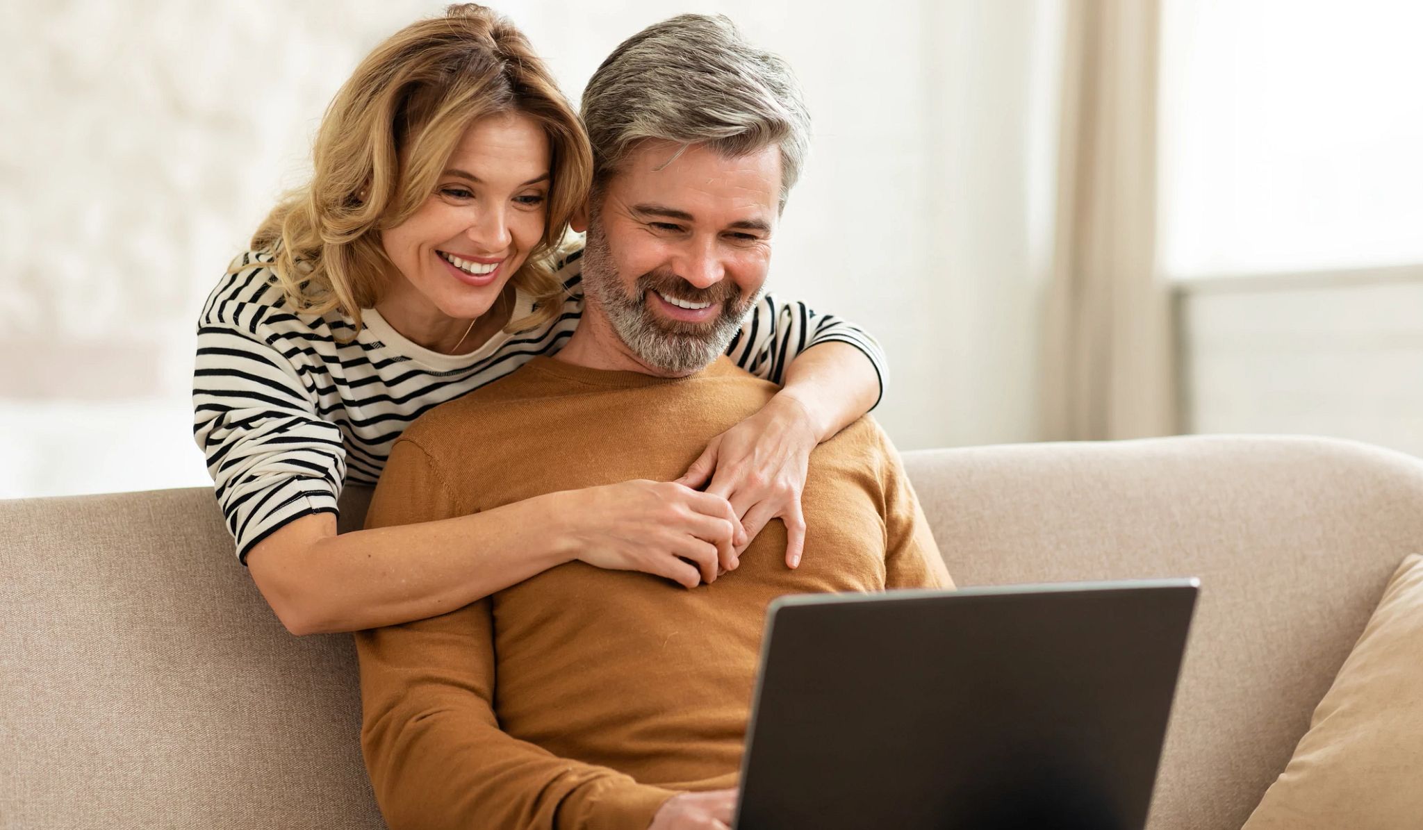 Happy Middle Aged Couple Using Laptop Computer Browsing Internet At Home. Wife Hugging Husband While Man orking Online Sitting On Sofa Indoor. Family Lifestyle And Technology Concept Happy Middle Aged Couple Using Laptop Computer Browsing Internet At Home. Wife Hugging Husband While Man orking Online Sitting On Sofa Indoor. Family Lifestyle And Technology Concept