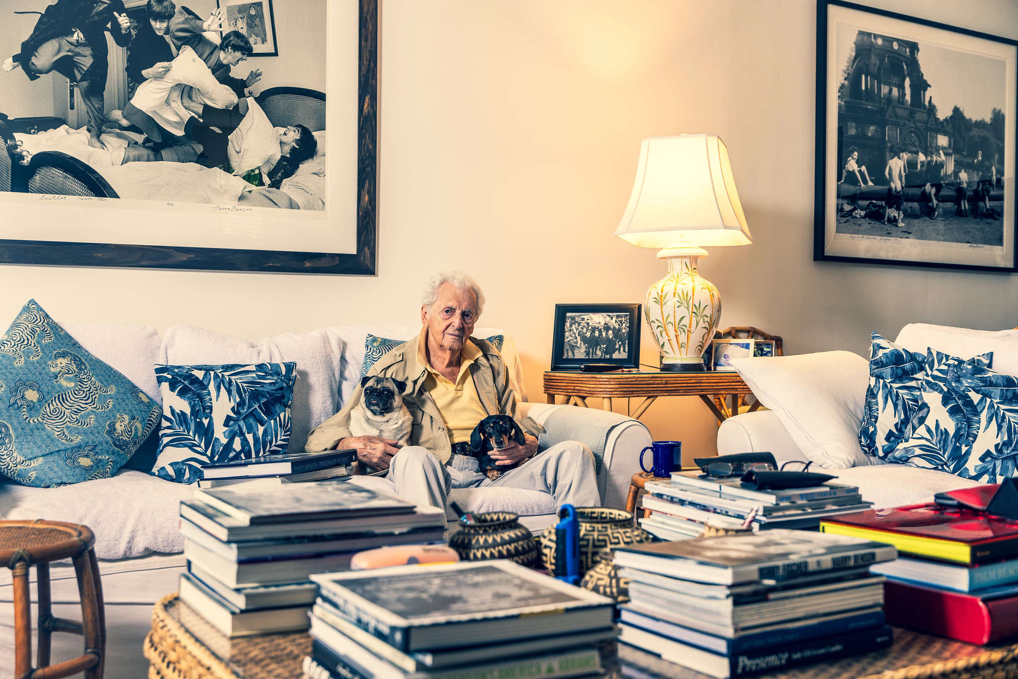 harry benson sits on a couch with two dogs on his lap. around him are dozens of books