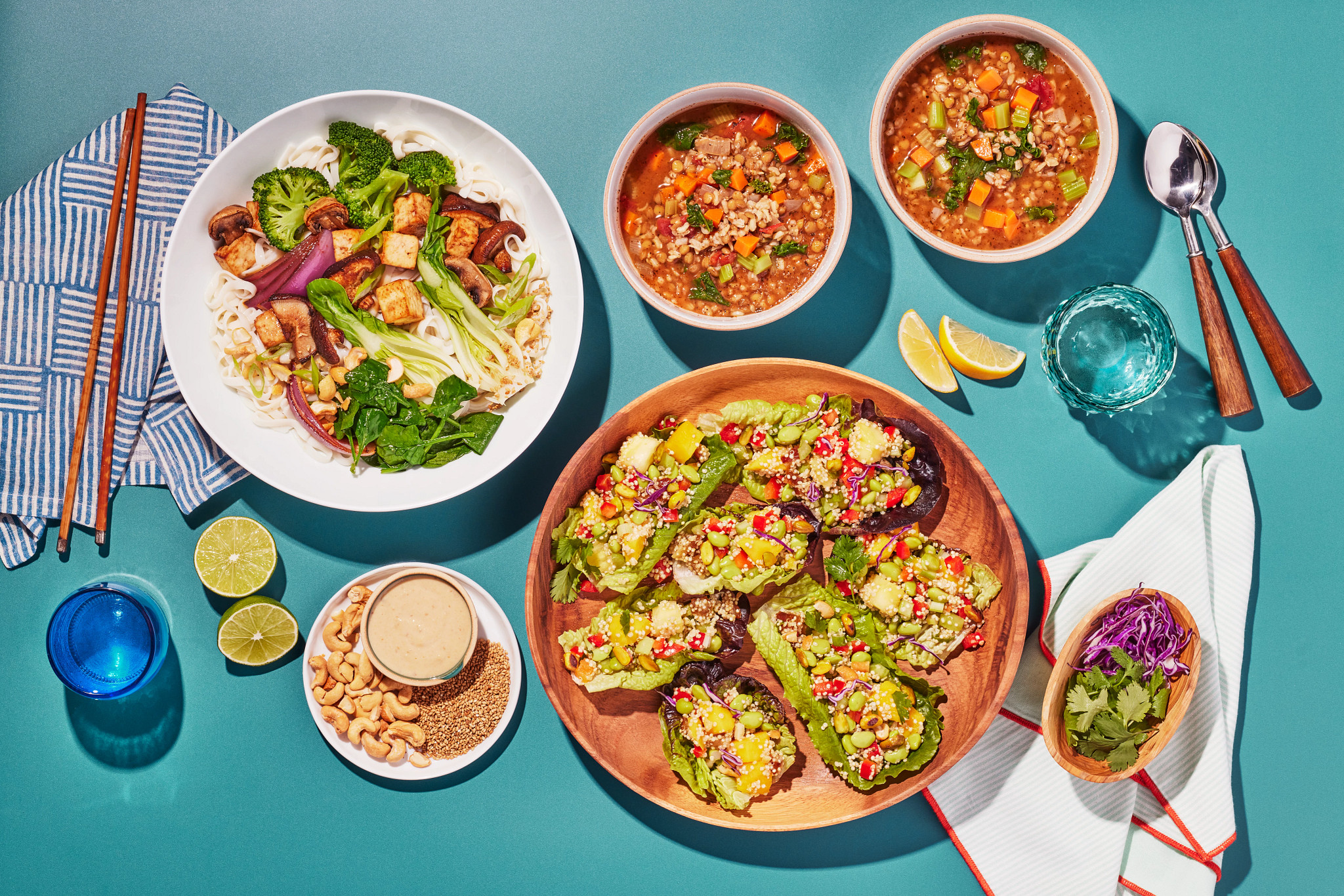Overhead photo of colorful plant-based meals including vegetable rice noodles with tofu and broccoli, hearty grain and vegetable soup, and lettuce wraps topped with quinoa, edamame, and fresh produce