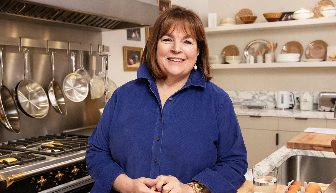 Ina Garten, chef, in blue long sleeve button up blouse, kitchen stove, white shelves, marble backsplash Ina Garten, chef, in blue long sleeve button up blouse, kitchen stove, white shelves, marble backsplash