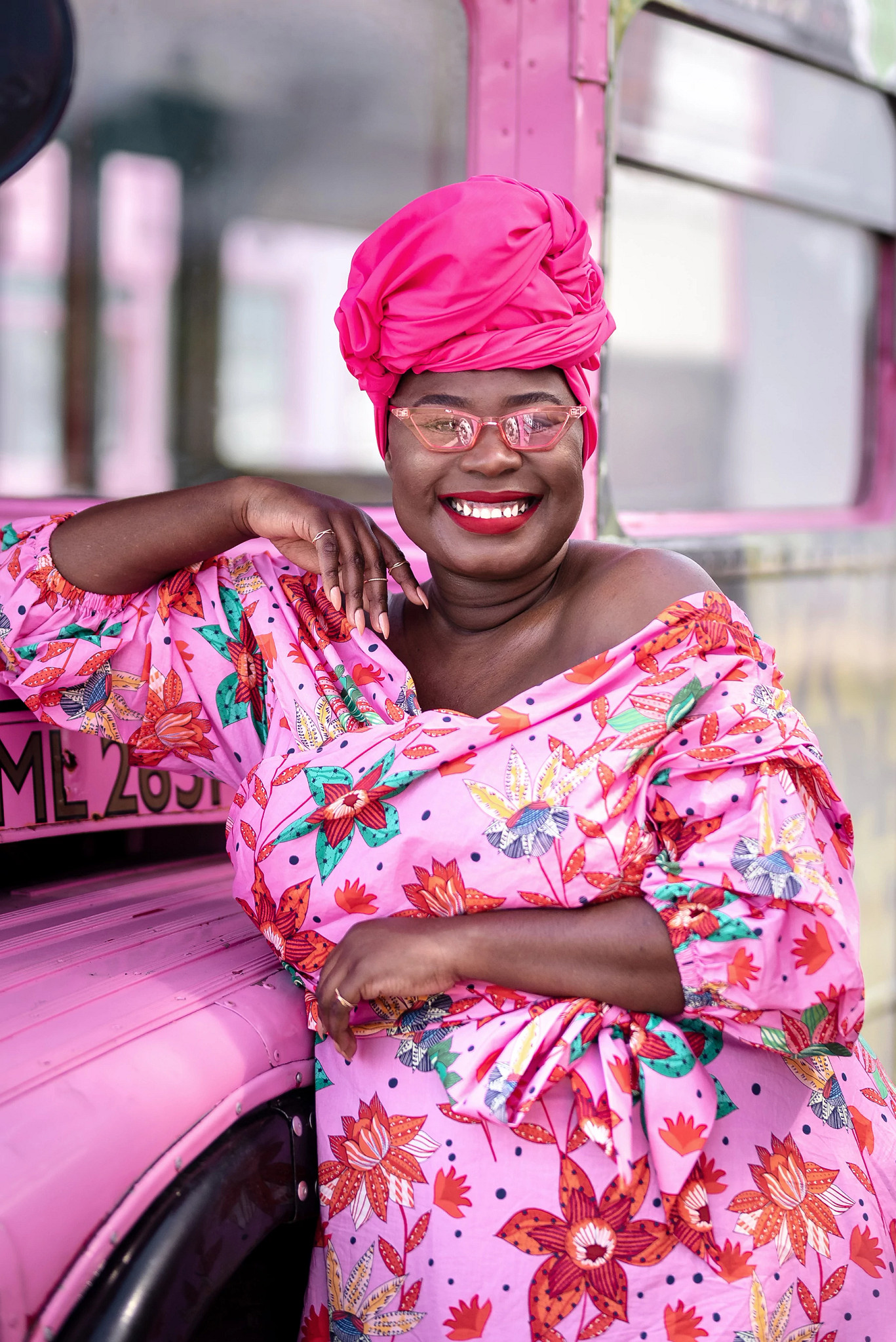 A person wearing a bright pink headwrap and floral pink dress poses beside a pink vehicle