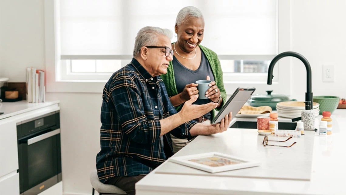 older man sitting at kitchen island in front of sink holding a tablet and looking at it with a woman standing next to him holding a coffee mug and smiling