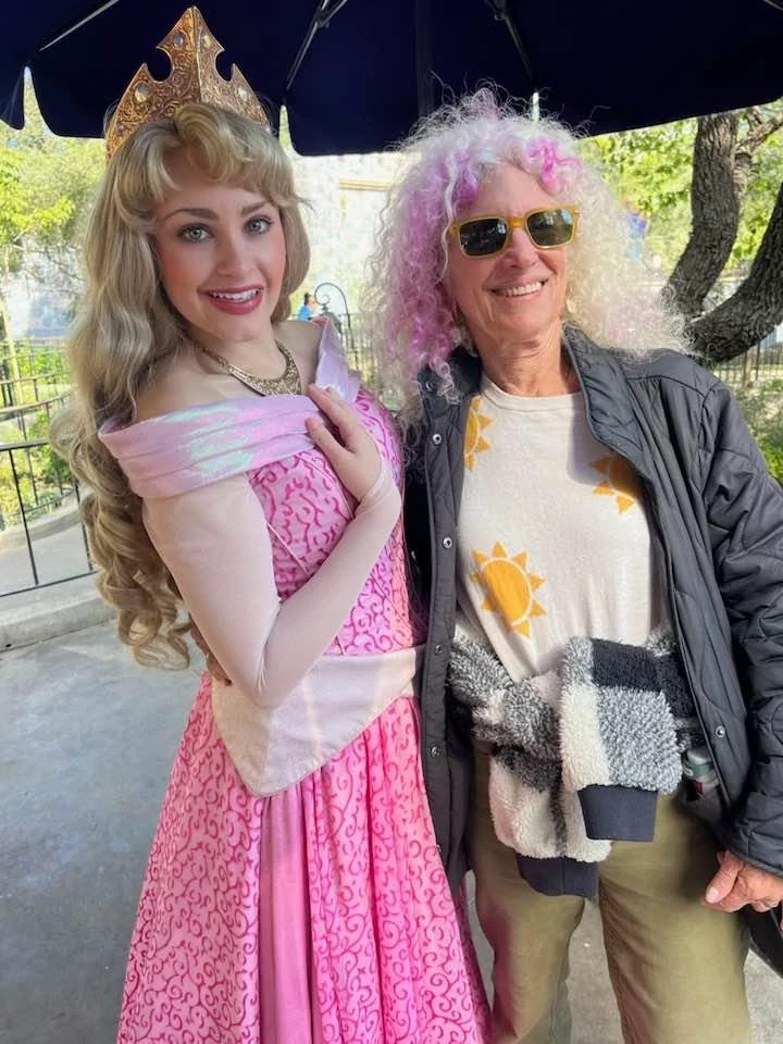 a woman with pink-streaked white hair smiles next to a woman dressed as sleeping beauty