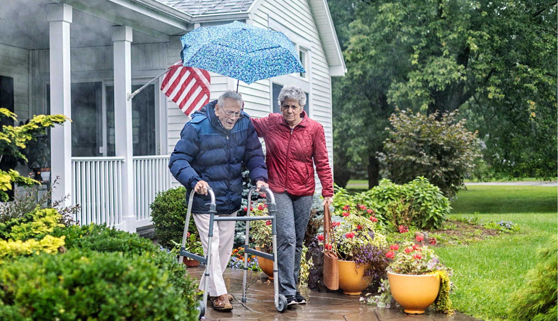Congress Passes Bill to Help Veterans Age in Place a caregiver holds an umbrella over a man using walker as they exit a house