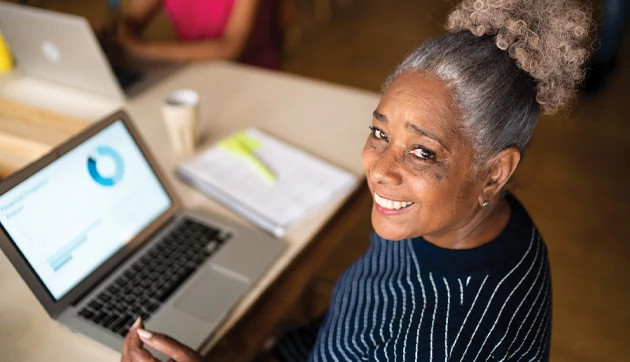 A smiling woman looks up while working on her laptop.