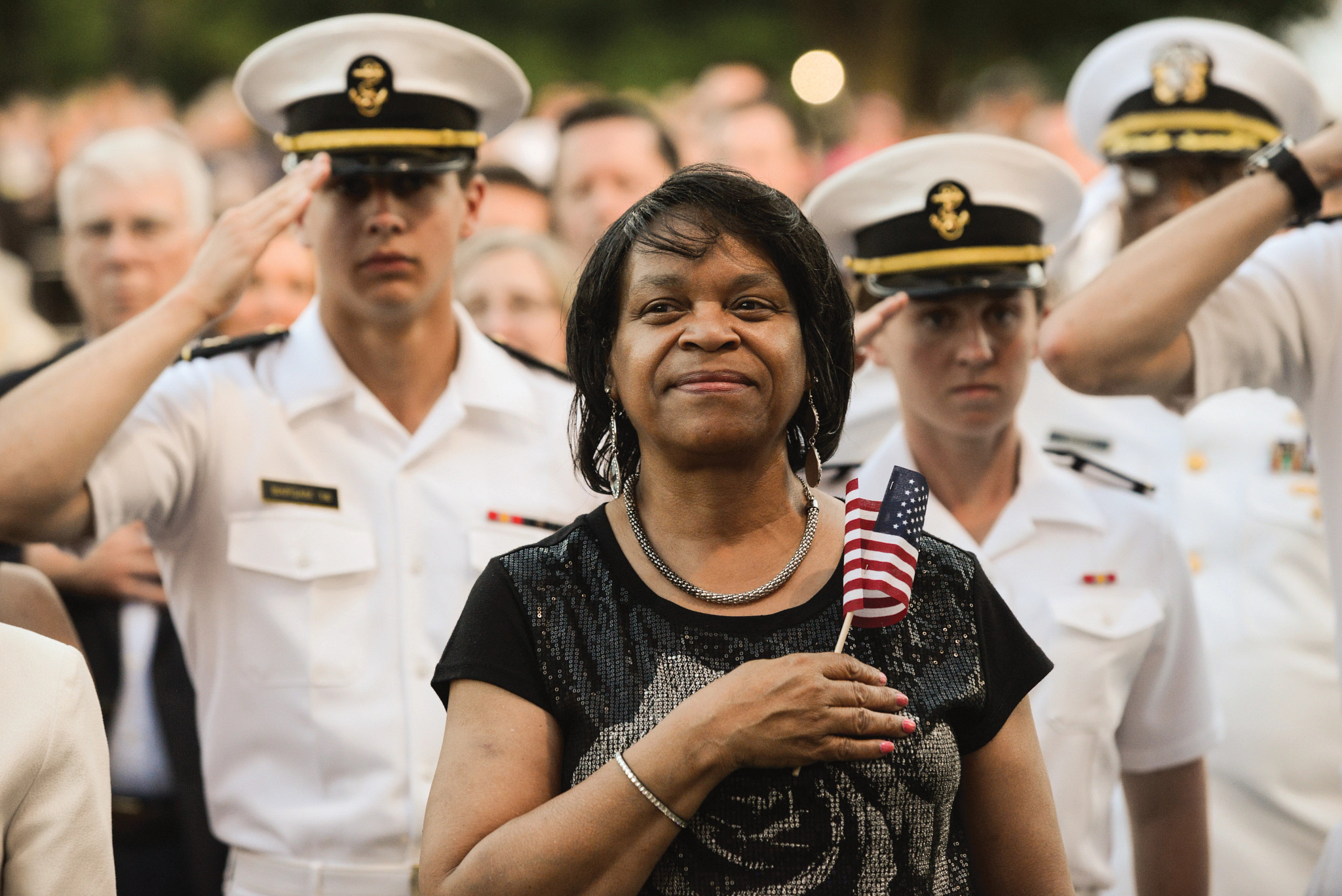 a person smiling while holding a miniature american flag over their heart while u s military service members in uniform salute in the background