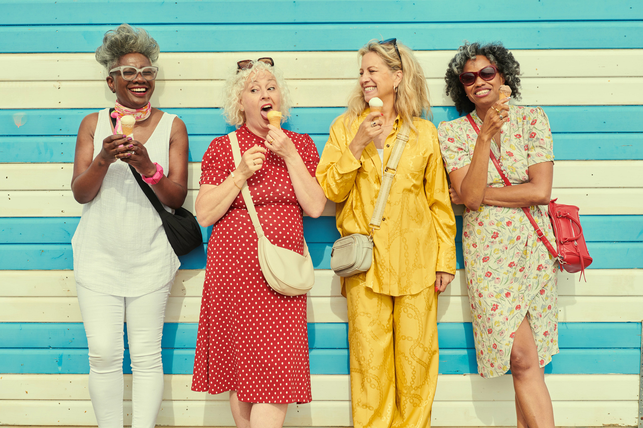 Four smiling older adult women laughing and eating ice cream cones while leaning against a blue and white striped wall.