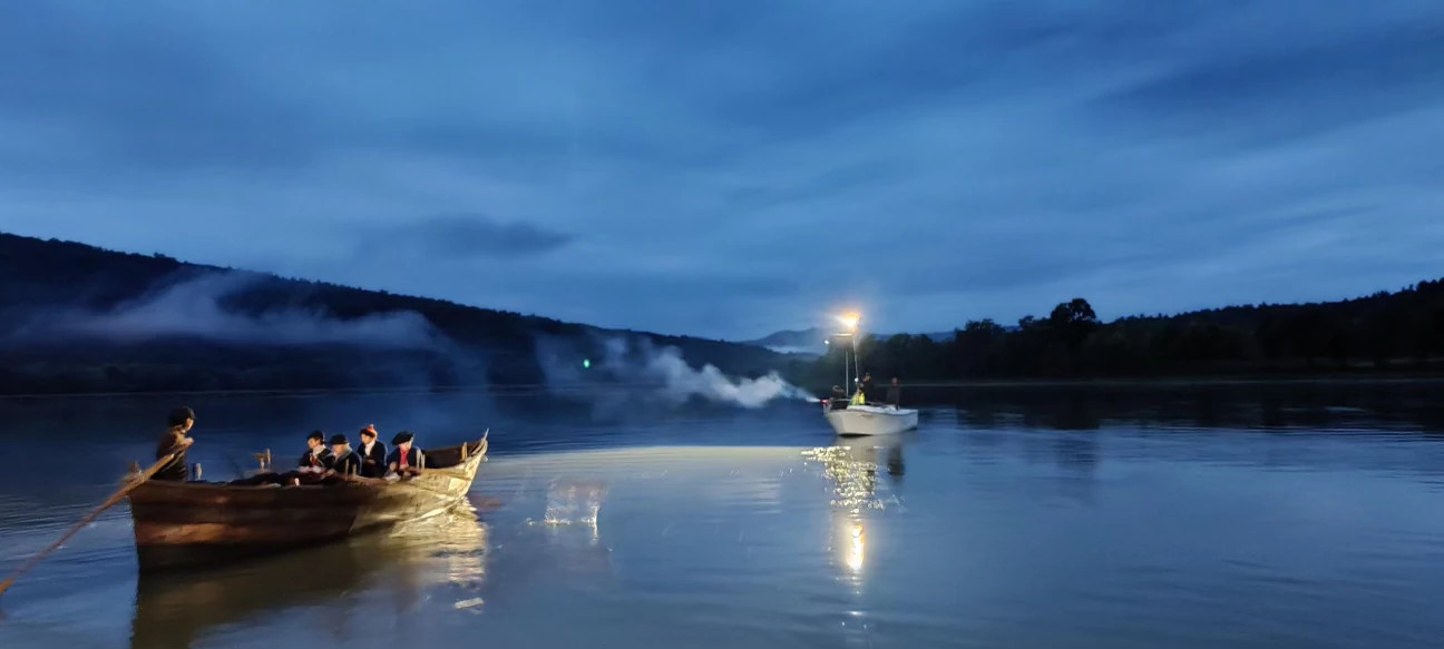 people dressed as soldiers in the american revolution rowing a wooden boat at night, followed by a modern boat with lights on it