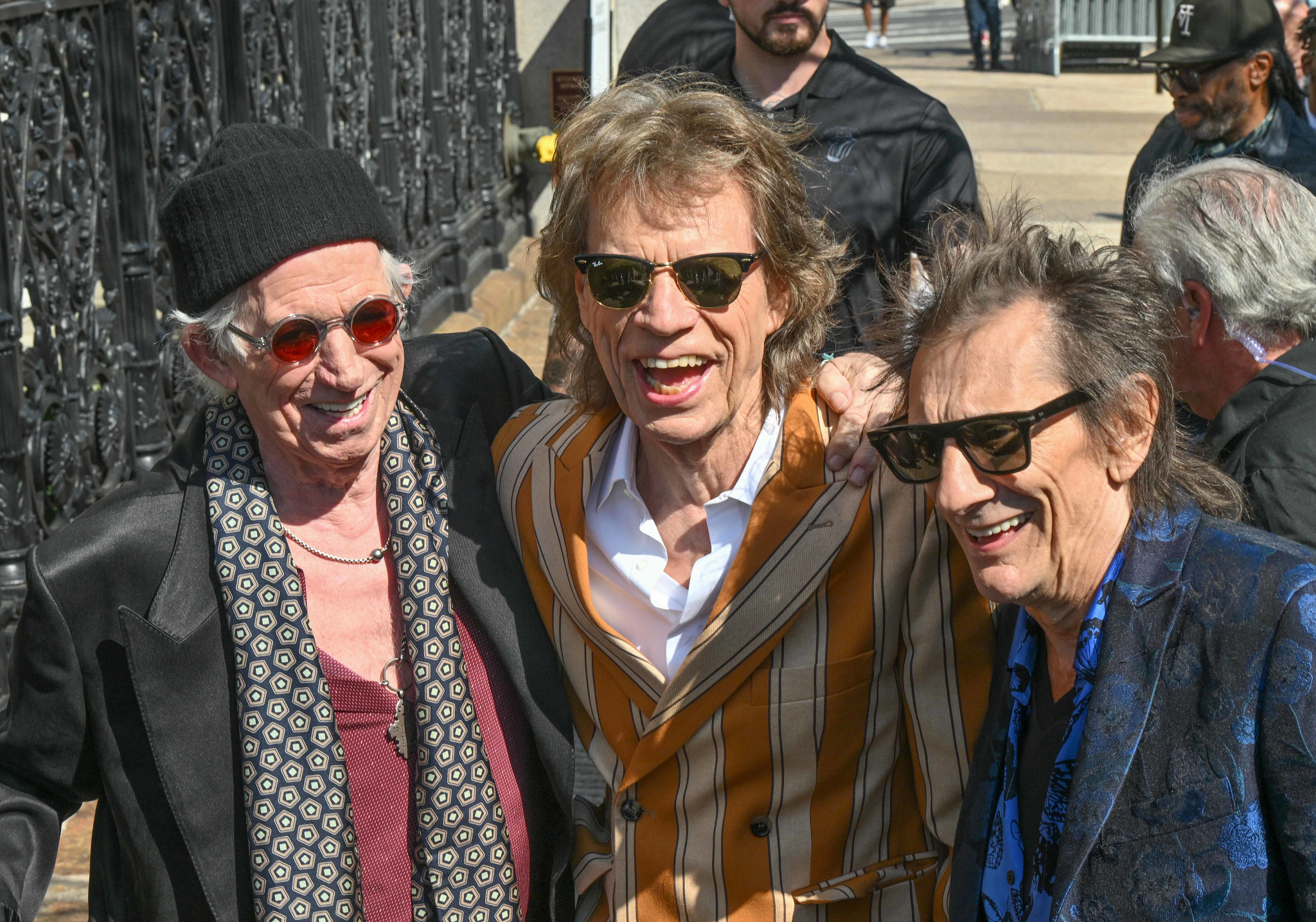 A candid outdoor photograph of Keith Richards, Mick Jagger, and Ronnie Wood smiling and laughing together. On the left, Keith Richards wears a black beanie, round red sunglasses, and a patterned scarf. In the center, Mick Jagger wears dark sunglasses and a gold-striped suit jacket. On the right, Ronnie Wood wears sunglasses and a blue brocade blazer.