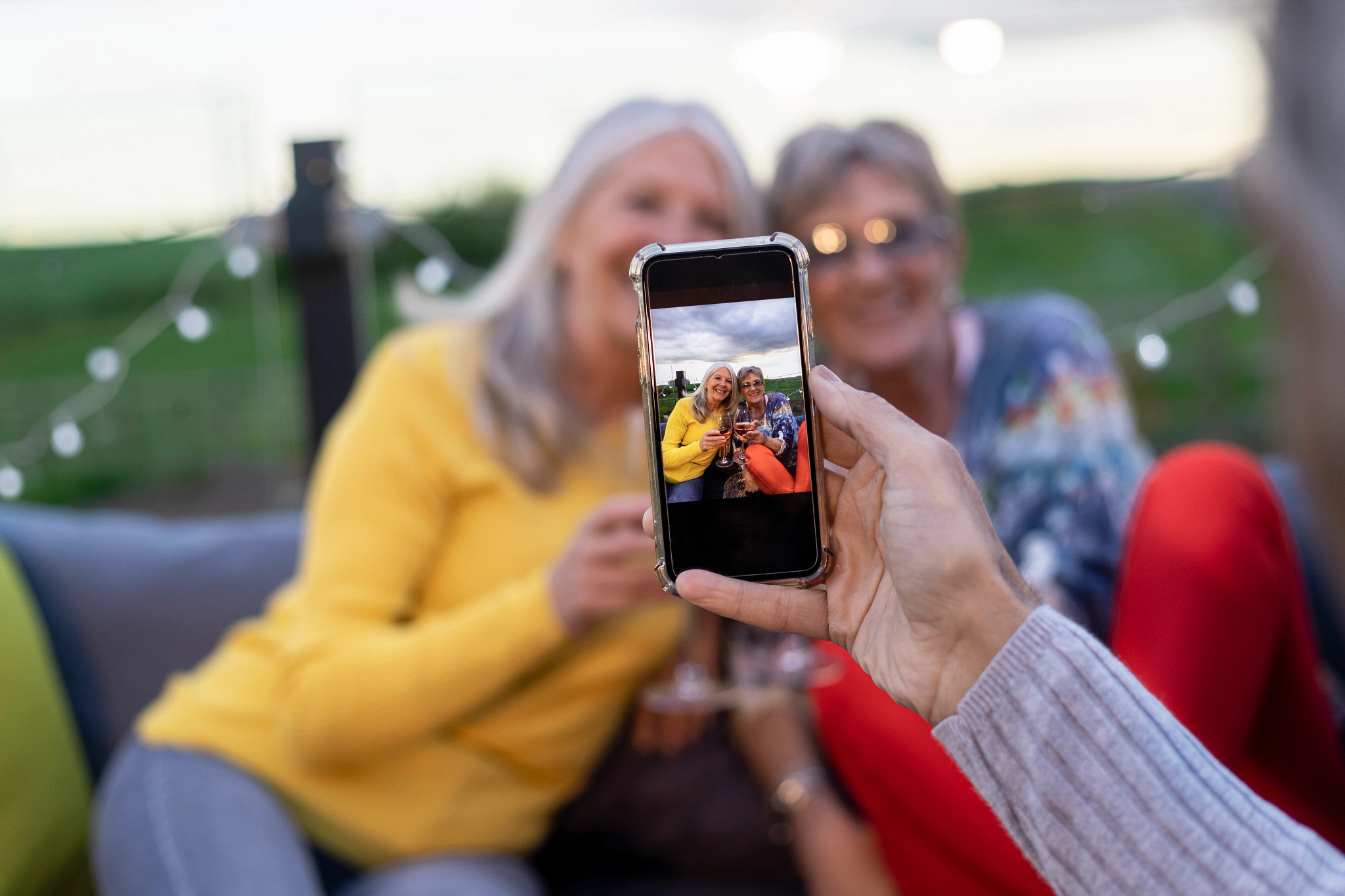Point of view shot of senior women getting their photo taken on a mobile phone sitting outside together drinking wine at a social gathering getting a photo together.