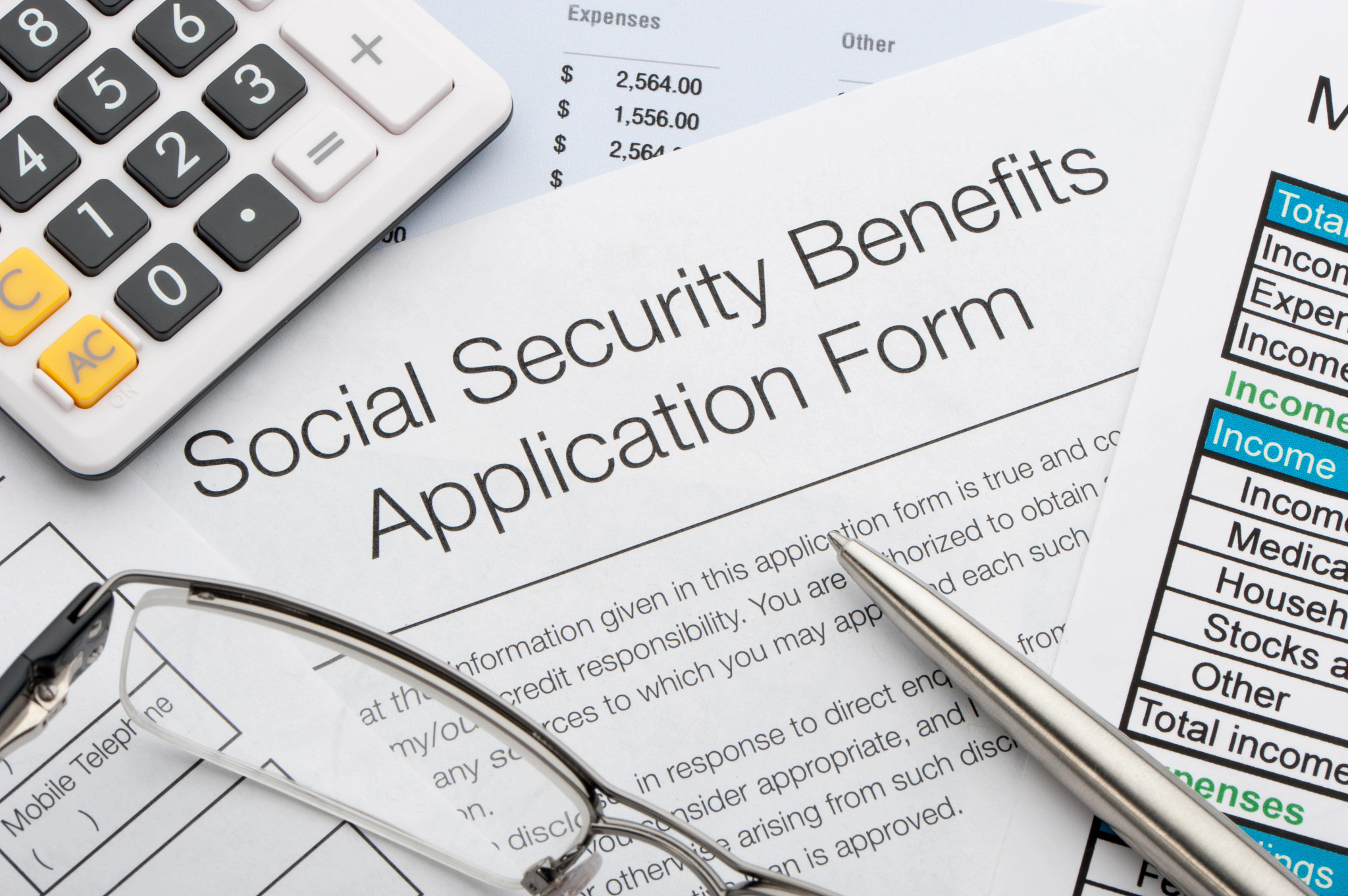 A top down view of a social security benefits application form with reading glasses, a pen, and a calculator sitting on the form.