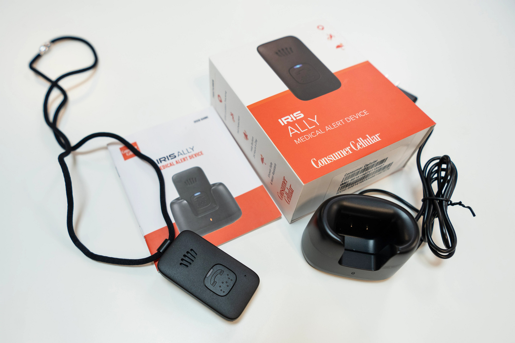 The IRIS Ally medical alert system on a table next to the charging port, manual and packaging