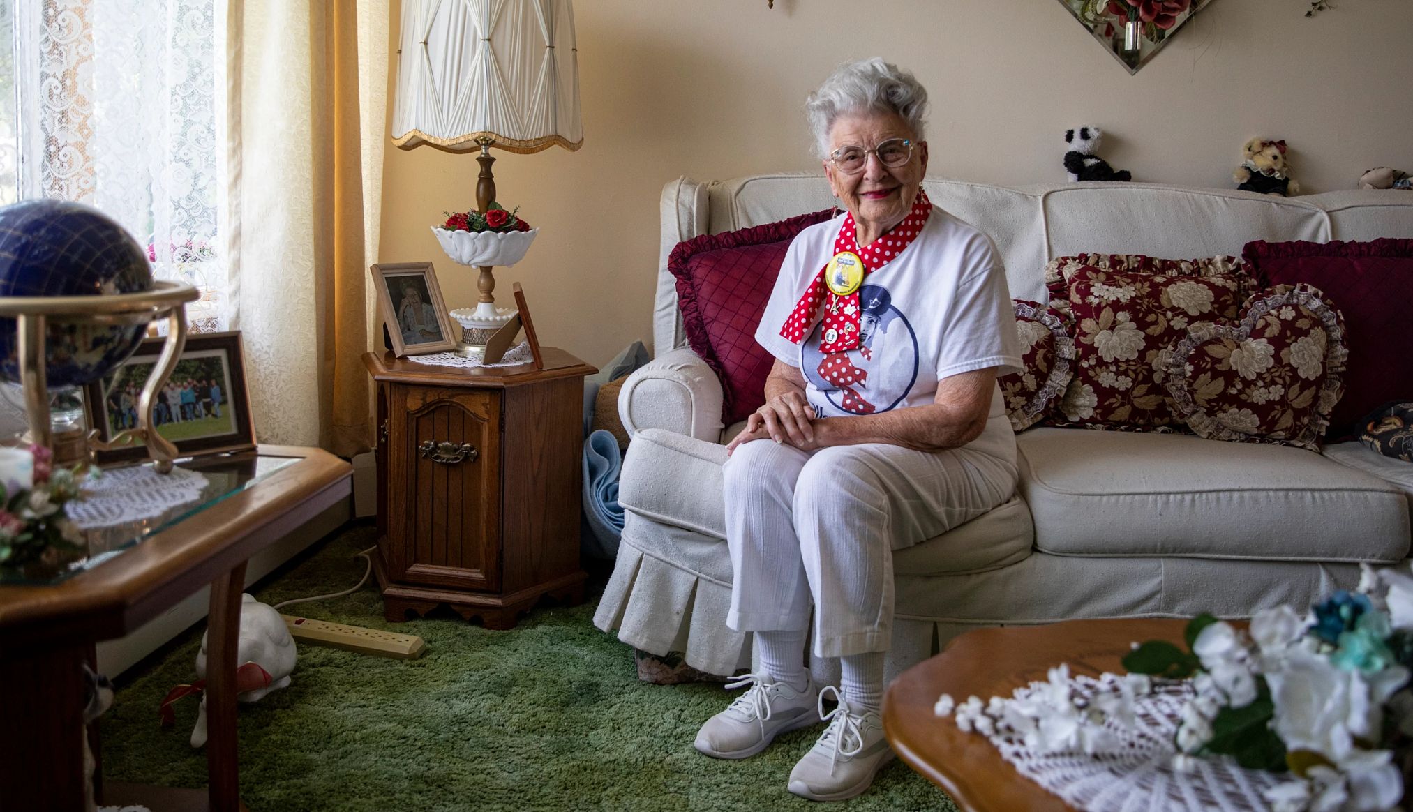 Mae Krier on Rosie the Riveter Mae Krier, wearing light colored clothes and a bright red and white polka dot scarf, sits on a couch in her living room.