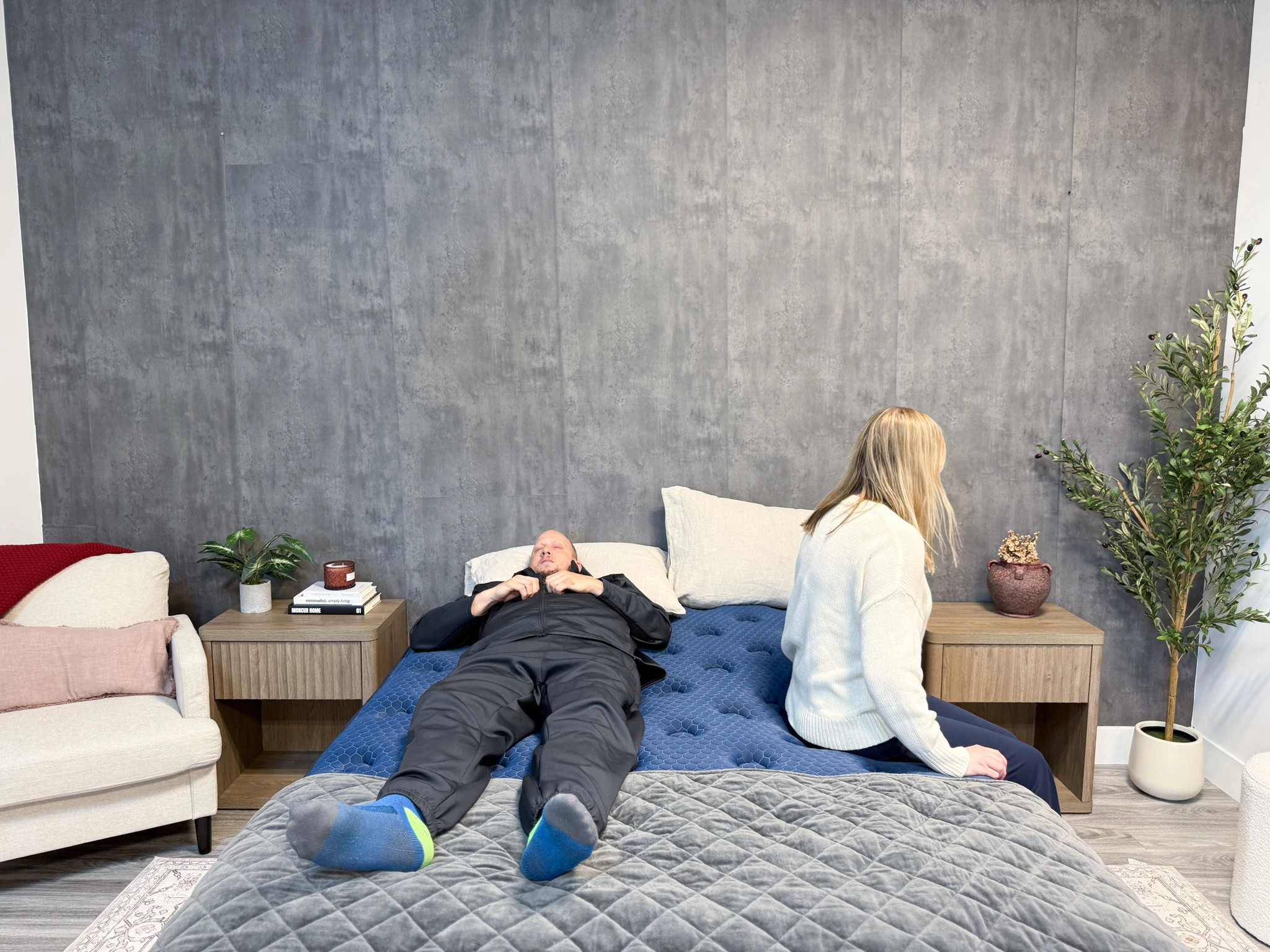  A woman sitting on the edge of a mattress while a man lies on his back on the other side