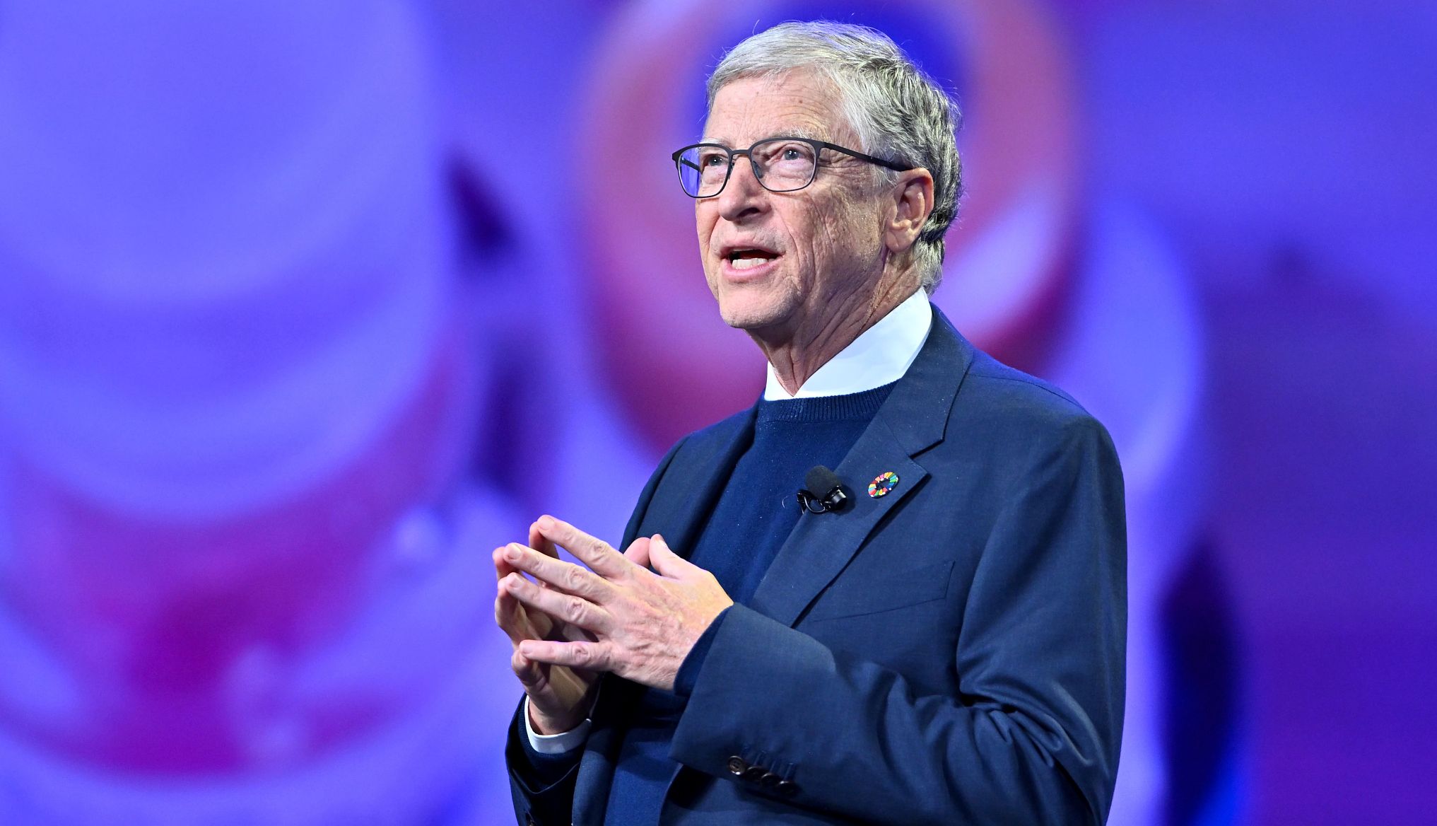 Bill Gates Motivational Speech Bill Gates is giving a speech at an event, wearing a navy blue suit, sweater, and a white shirt