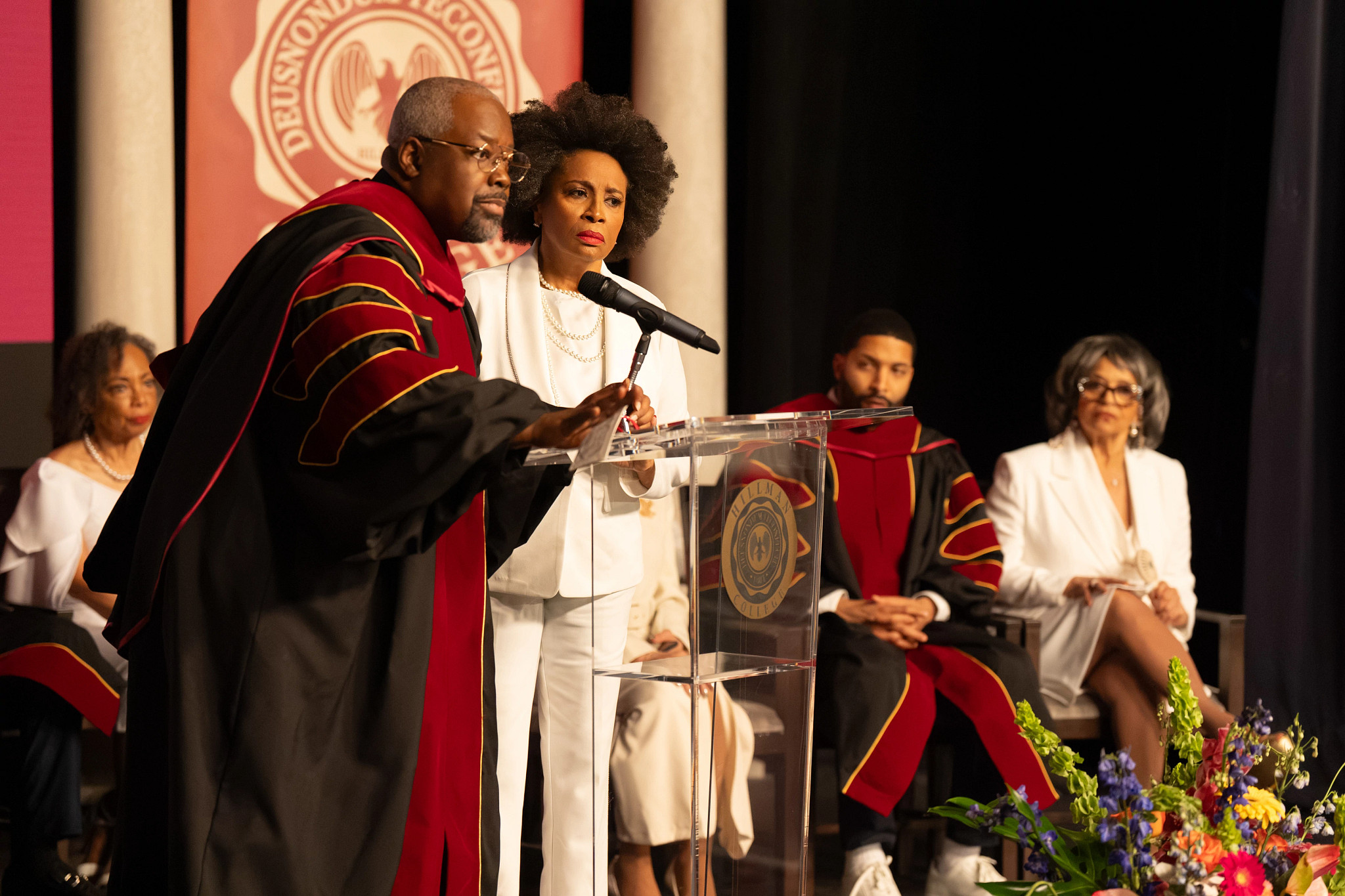 Kadeem Hardison and Jenifer Lewis stand at a clear podium during a formal academic ceremony with participants in ceremonial robes seated on stage in A Different World sequel series