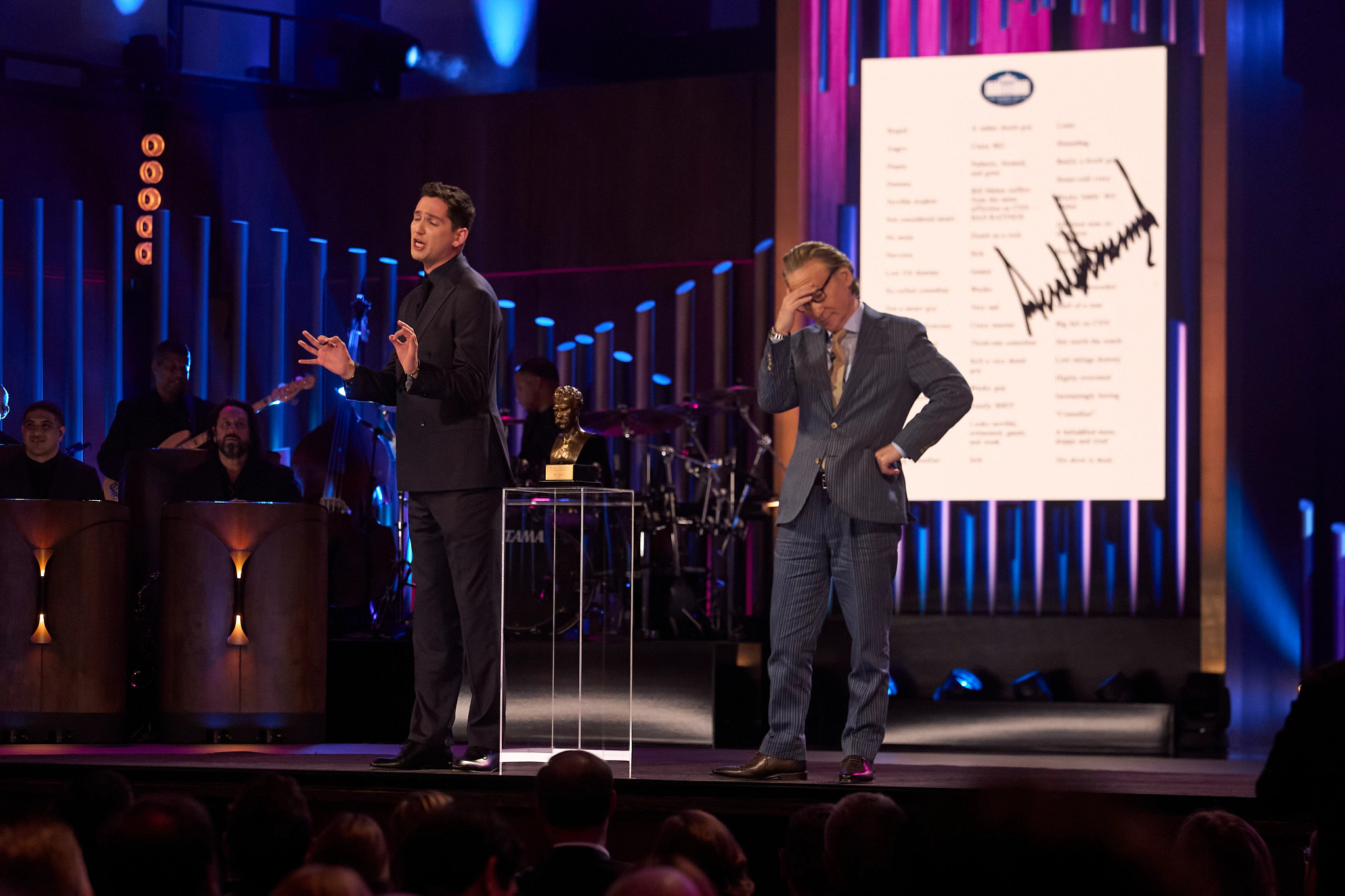matt friend stands on the stage at the mark twain prize award ceremony in front of an exasperated looking bill maher