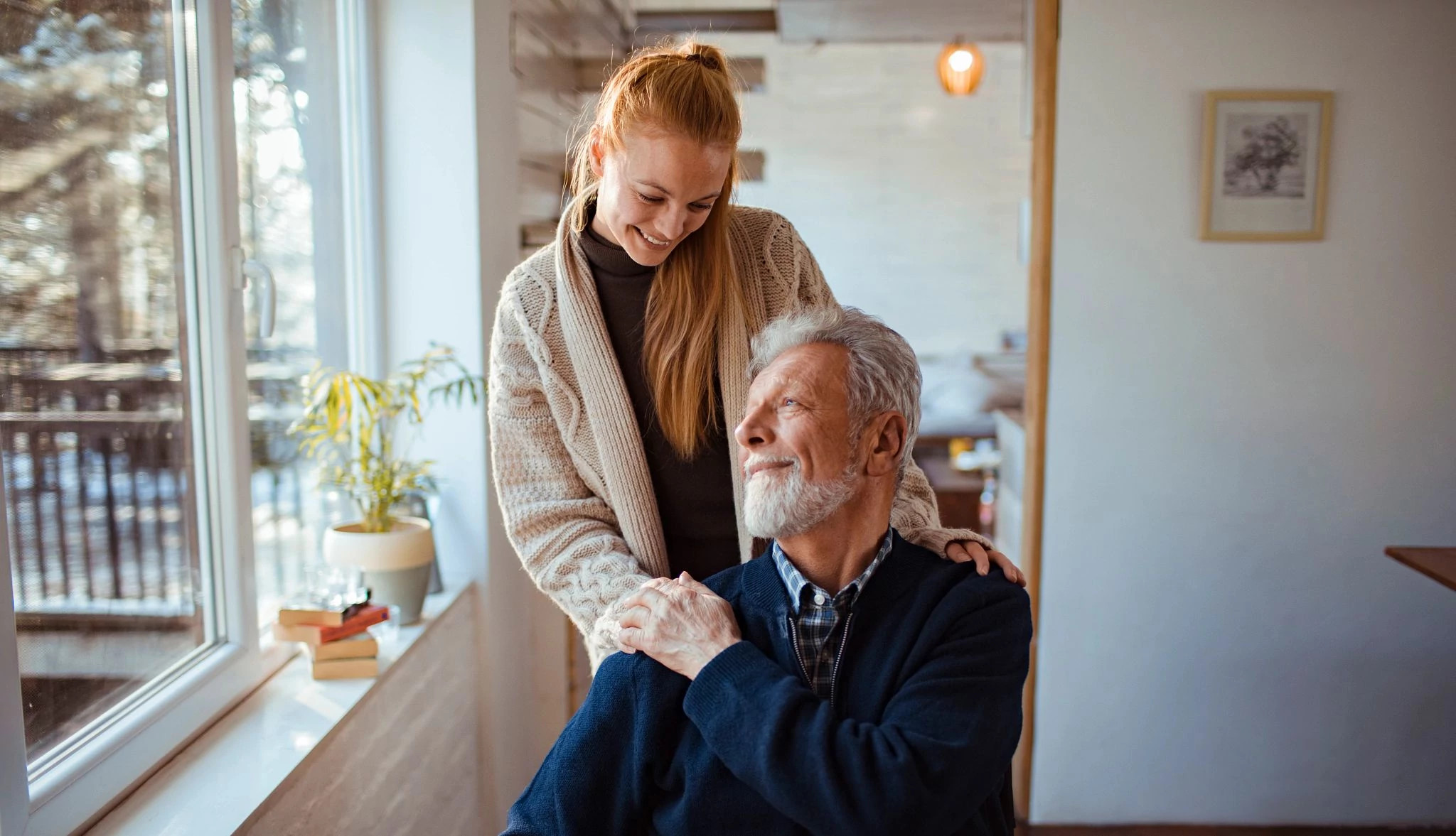 A young woman stands behind an older man sitting in a chair near a window, gently resting a supportive hand on his shoulder as they talk