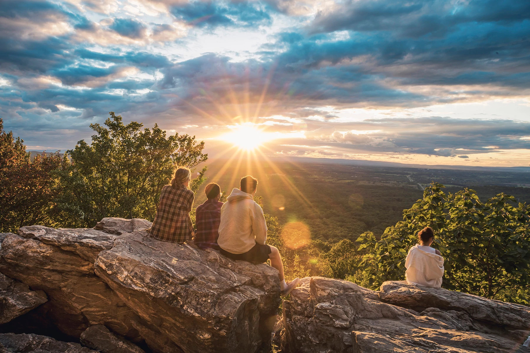 Four hikers sitting on a rocky overlook watching sunset across a forested valley