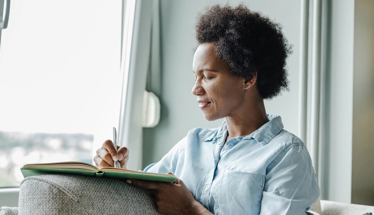 A woman sits on a couch near a window and writes in a notebook.