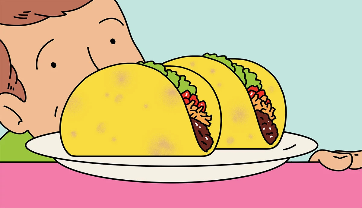 A person looking eagerly at a plate of two hard-shell tacos filled with cheese, meat, and vegetables.