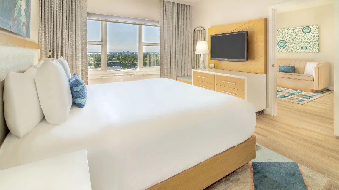 Radisson® bed with white linens in hotel room with window, tv, separate room with couch