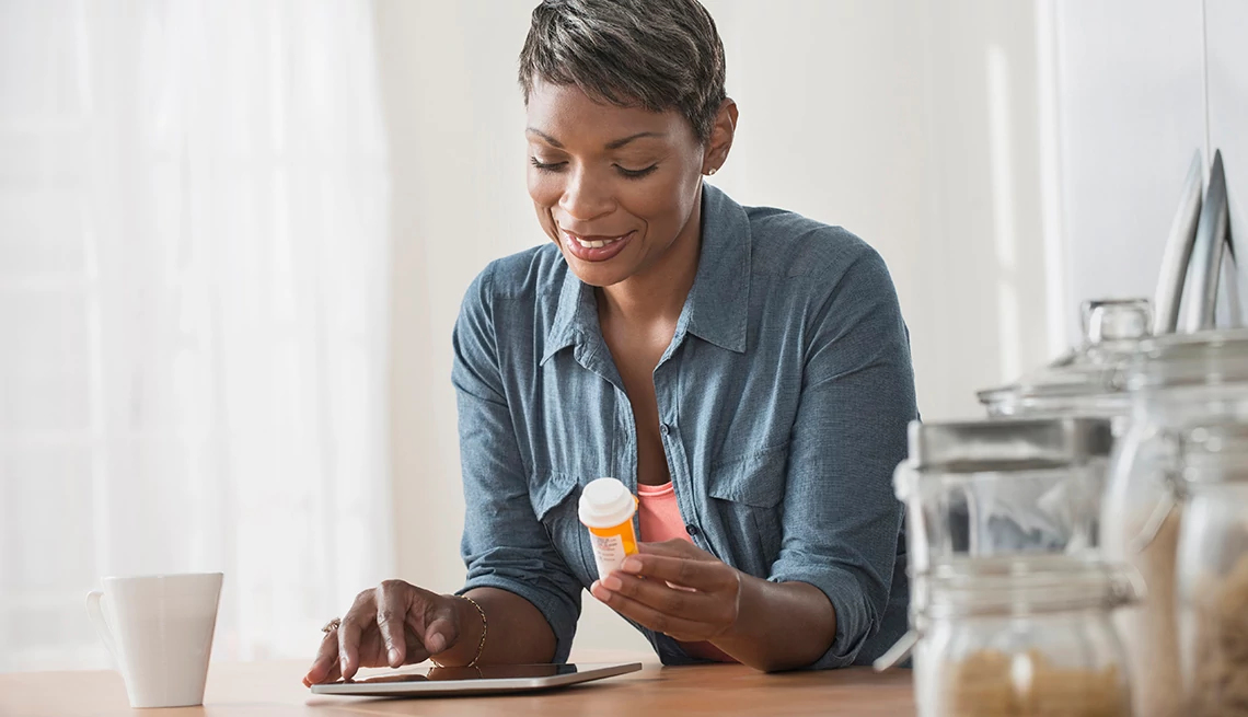 Una afroamericana sostiene en la mano izquierda un frasco de medicina mientras mira su tableta Una afroamericana sostiene en la mano izquierda un frasco de medicina mientras mira su tableta