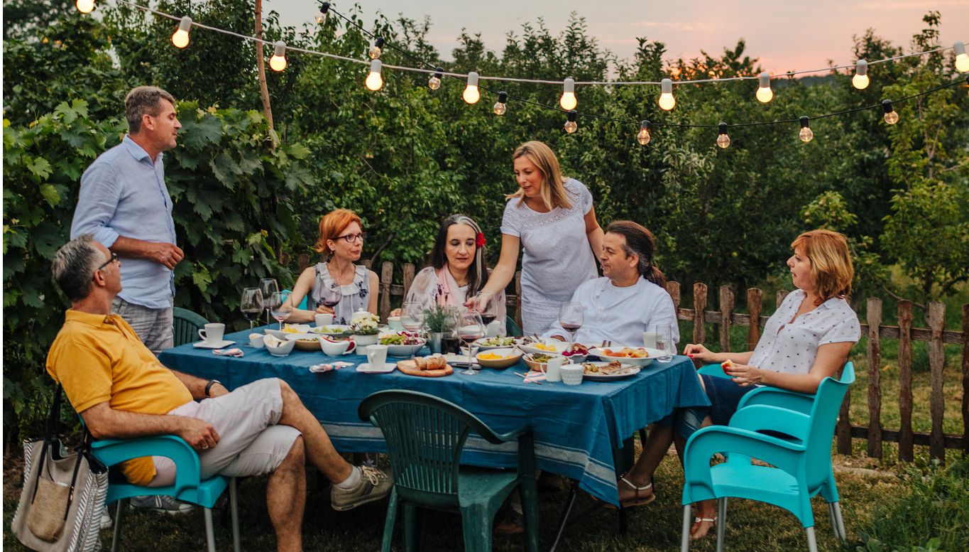 Group of people sitting and standing while drinking wine at night in front of a vineyard with string lights behind them. Group of people sitting and standing while drinking wine at night in front of a vineyard with string lights behind them.