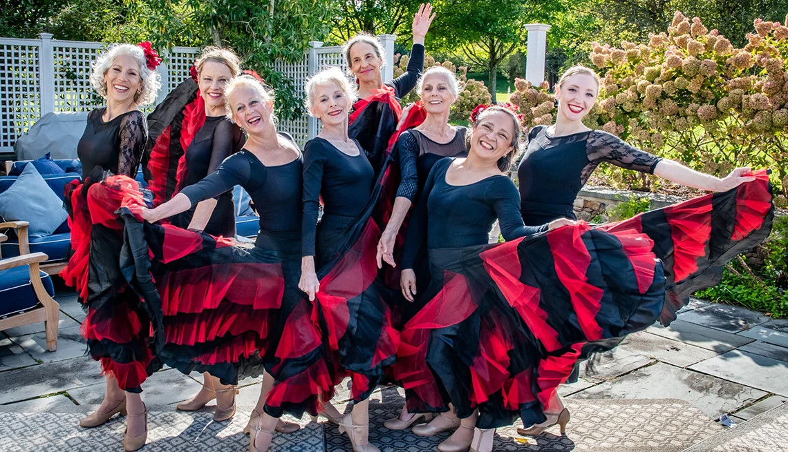 A group of eight smiling women in black dance leotards and vibrant red and black ruffled skirts pose outdoors, representing the Helping Hearts Through the Arts performers.