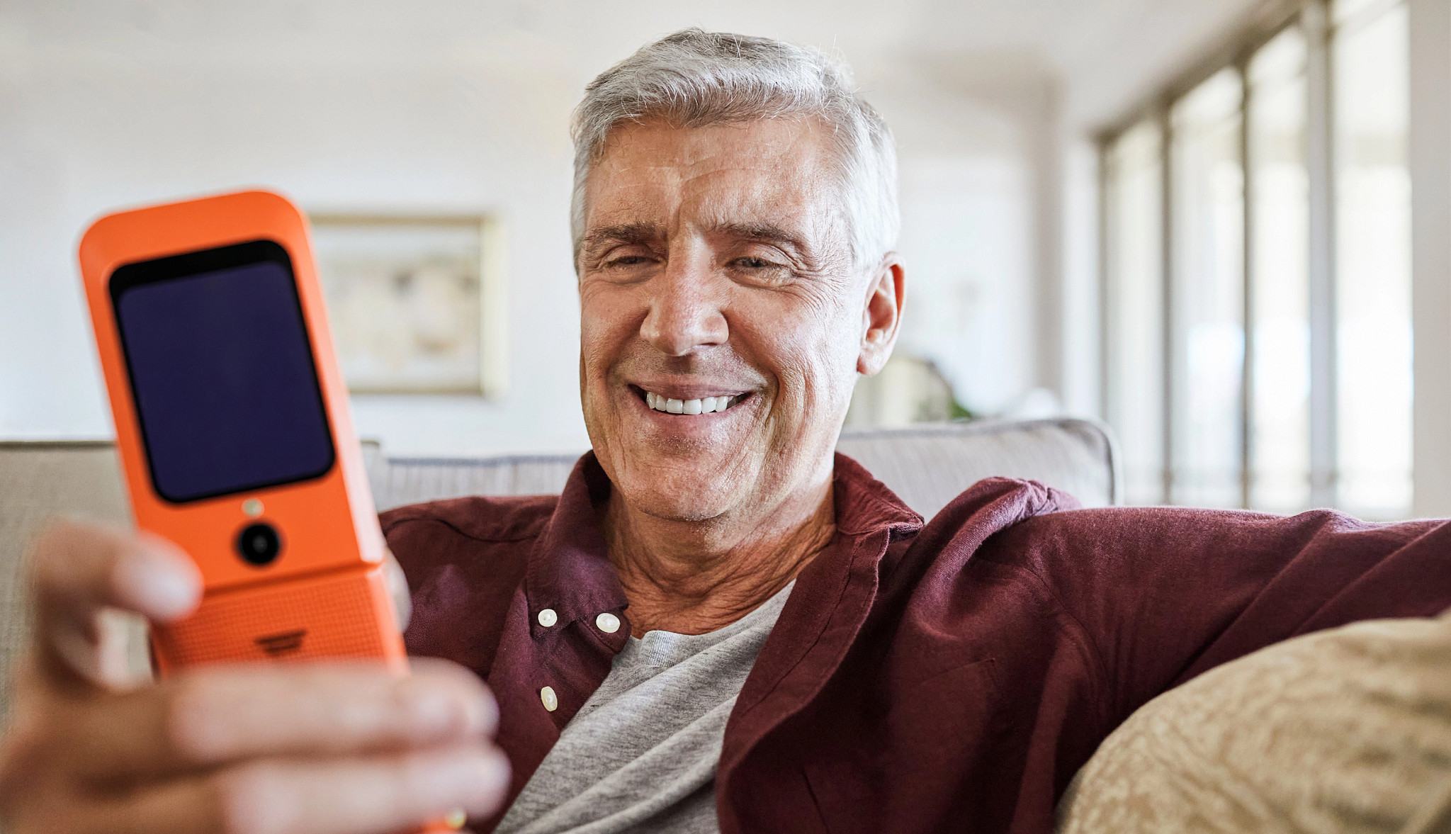 Mature man in a maroon shirt looking at his orange flip phone from Consumer Cellular