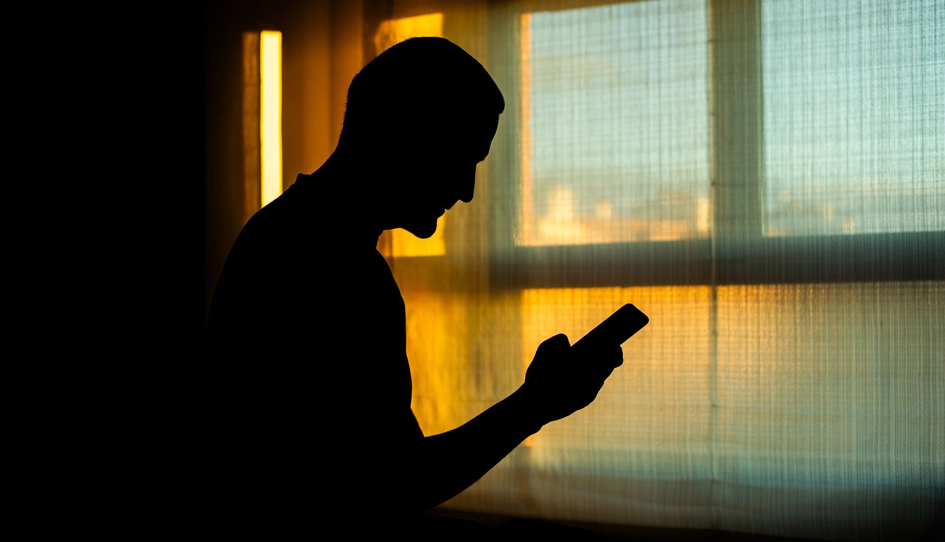 Social Security Drops Phone Cuts a man stands near a window, silhouetted by the setting sun. he is using a smartphone.
