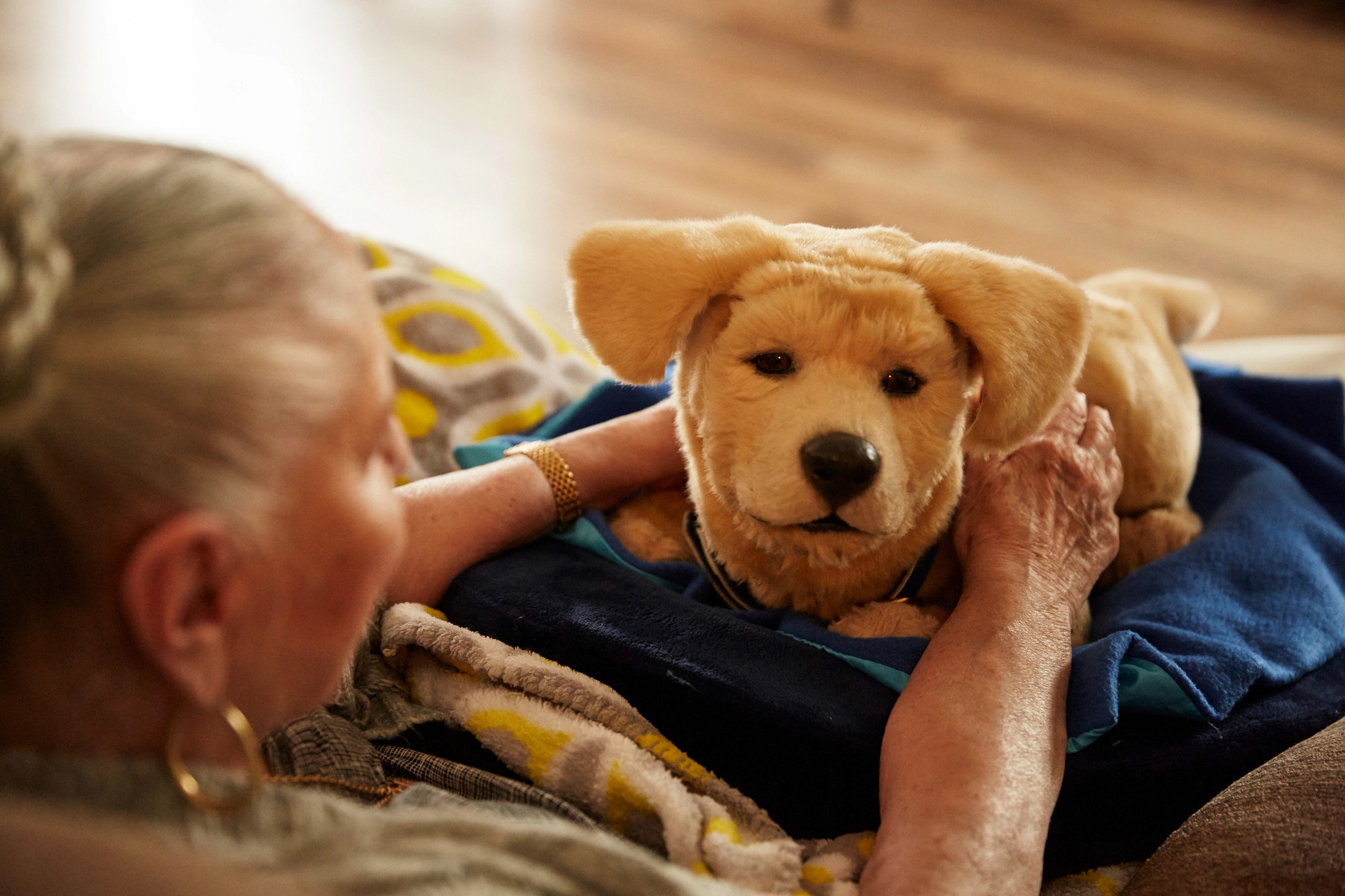 A photo shows an older adult woman with Tombot Jennie, a cuddly robot retriever