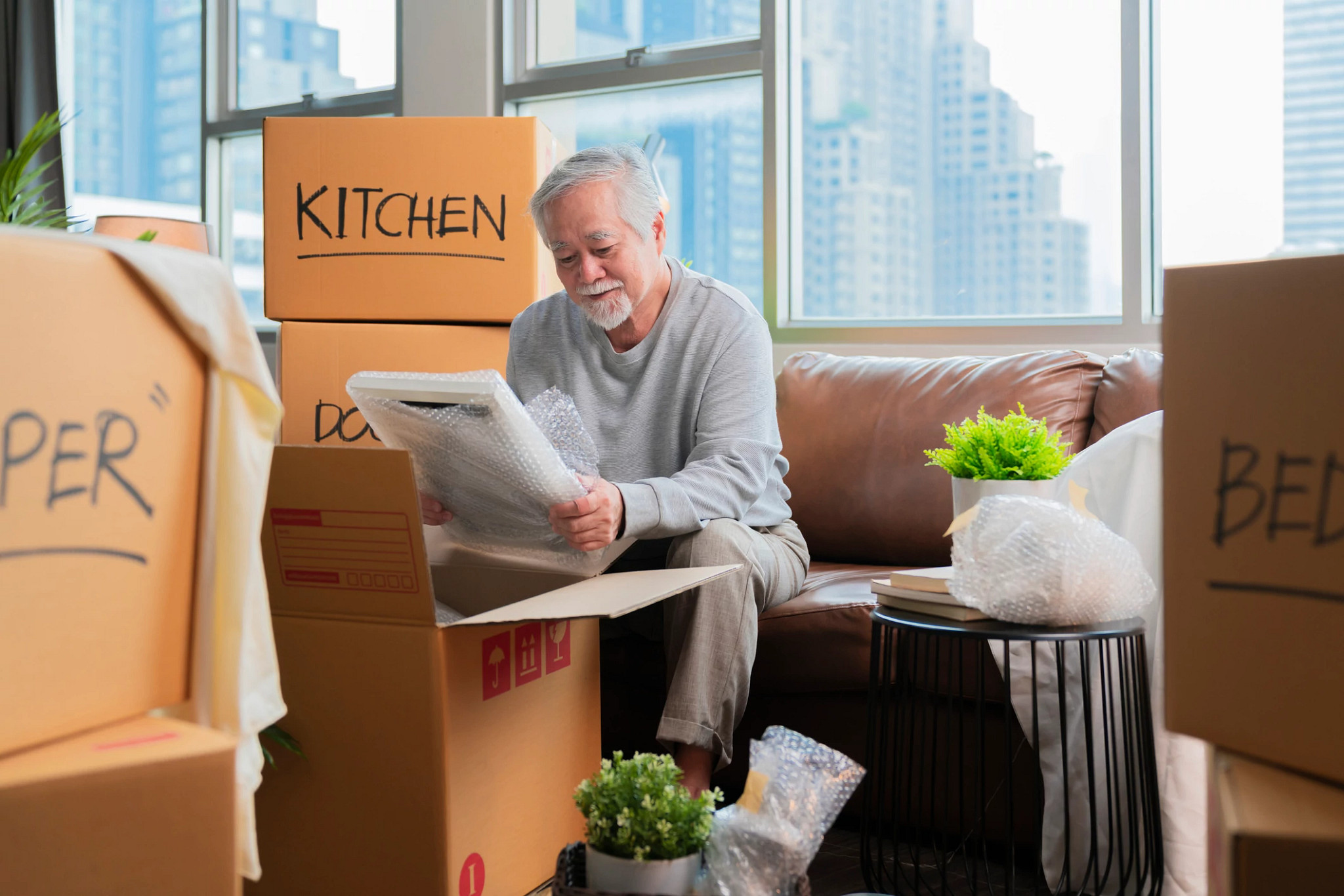 Horizontal medium shot of adult elder senior asian man male white beard sitting on floor preparing for moving to new house packing things into boxes and taping them in living room,home moving ideas concept
