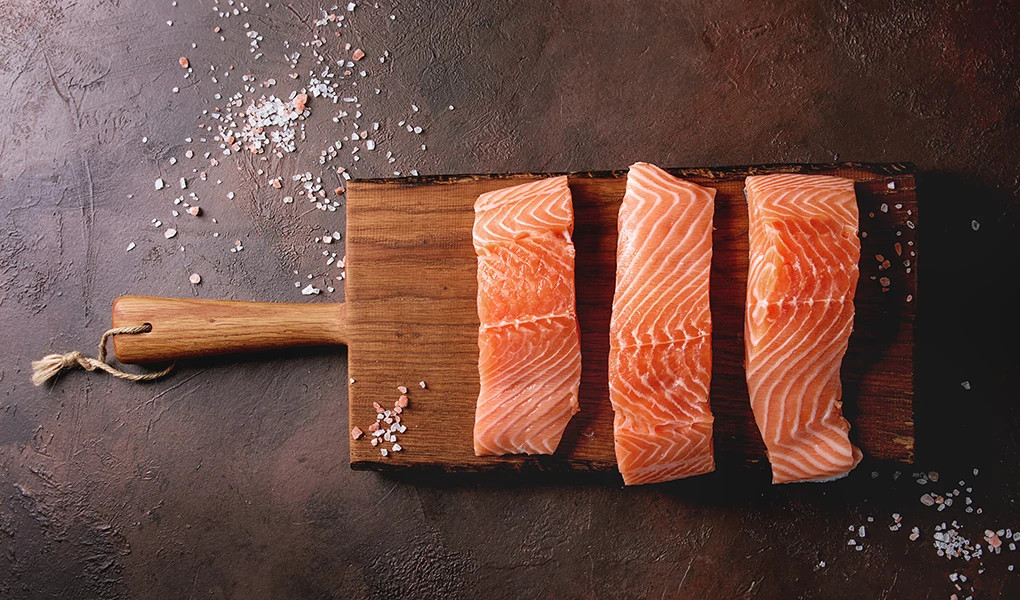 Sliced raw salmon on a wooden cutting board with sea salt and pepper against a dark brown background