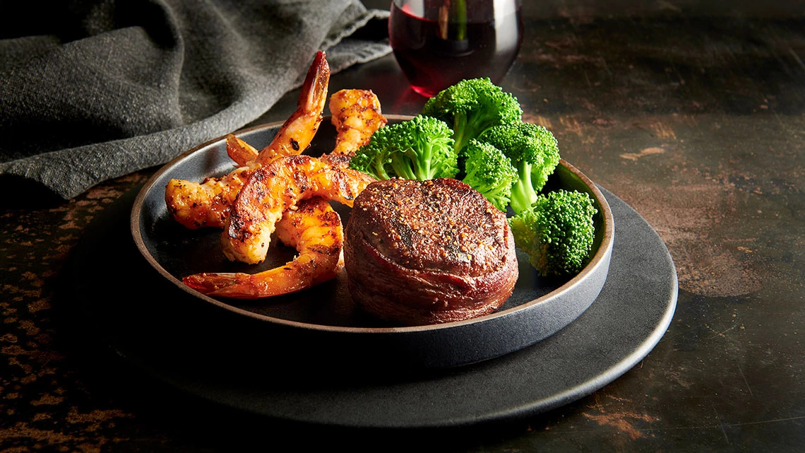 Saltgrass Steak House red glass wine next to a black plate of steak filet, shrimp, broccoli