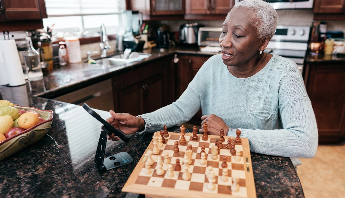 A woman plays chess over her phone while sitting in her kitchen.