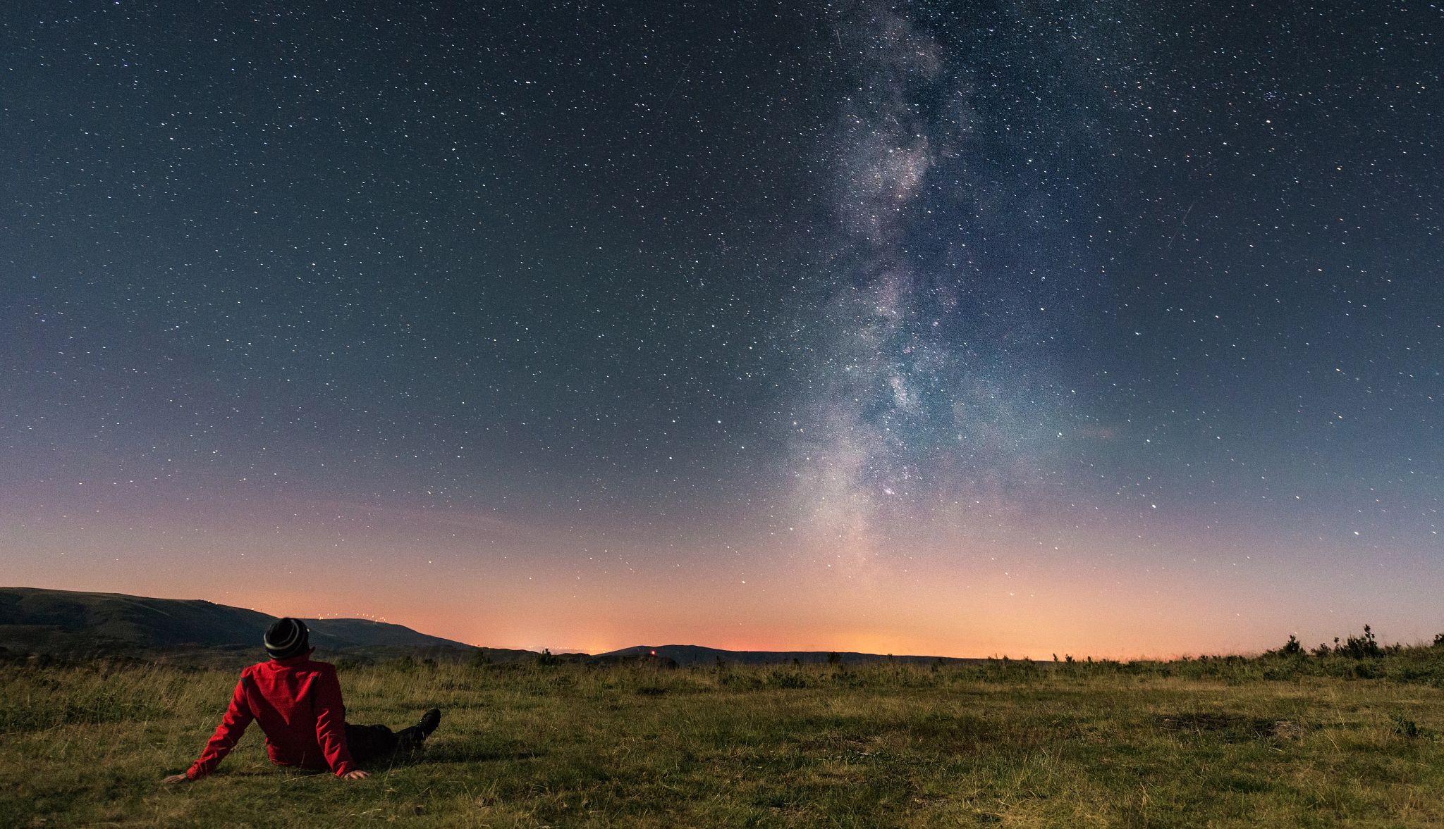 Image depicting the exploration of night sky A person sits amidst a field, gazing at the breathtaking Milky Way, a celestial river flowing across the night sky.