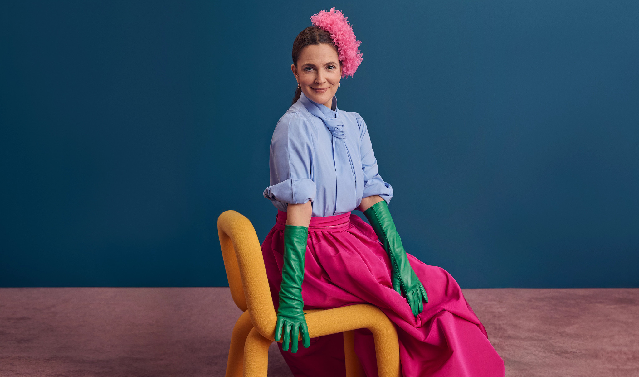 drew barrymore sits in a chair, wearing a blue shirt, bright pink skirt and hat, and green gloves