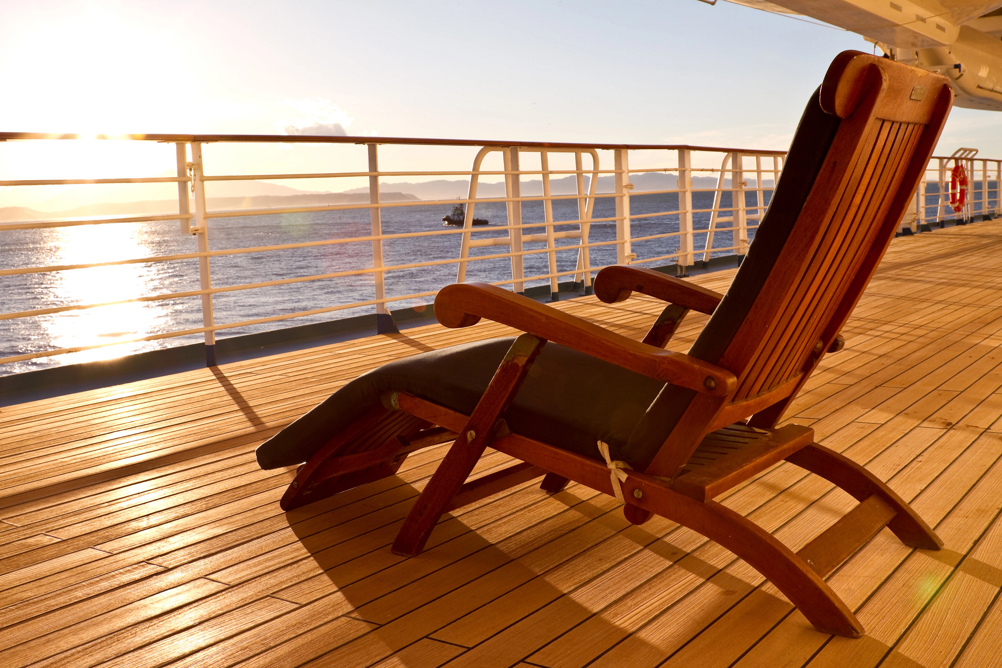 An empty wooden lounge chair faces the ocean on a cruise ship deck during golden-hour sunlight