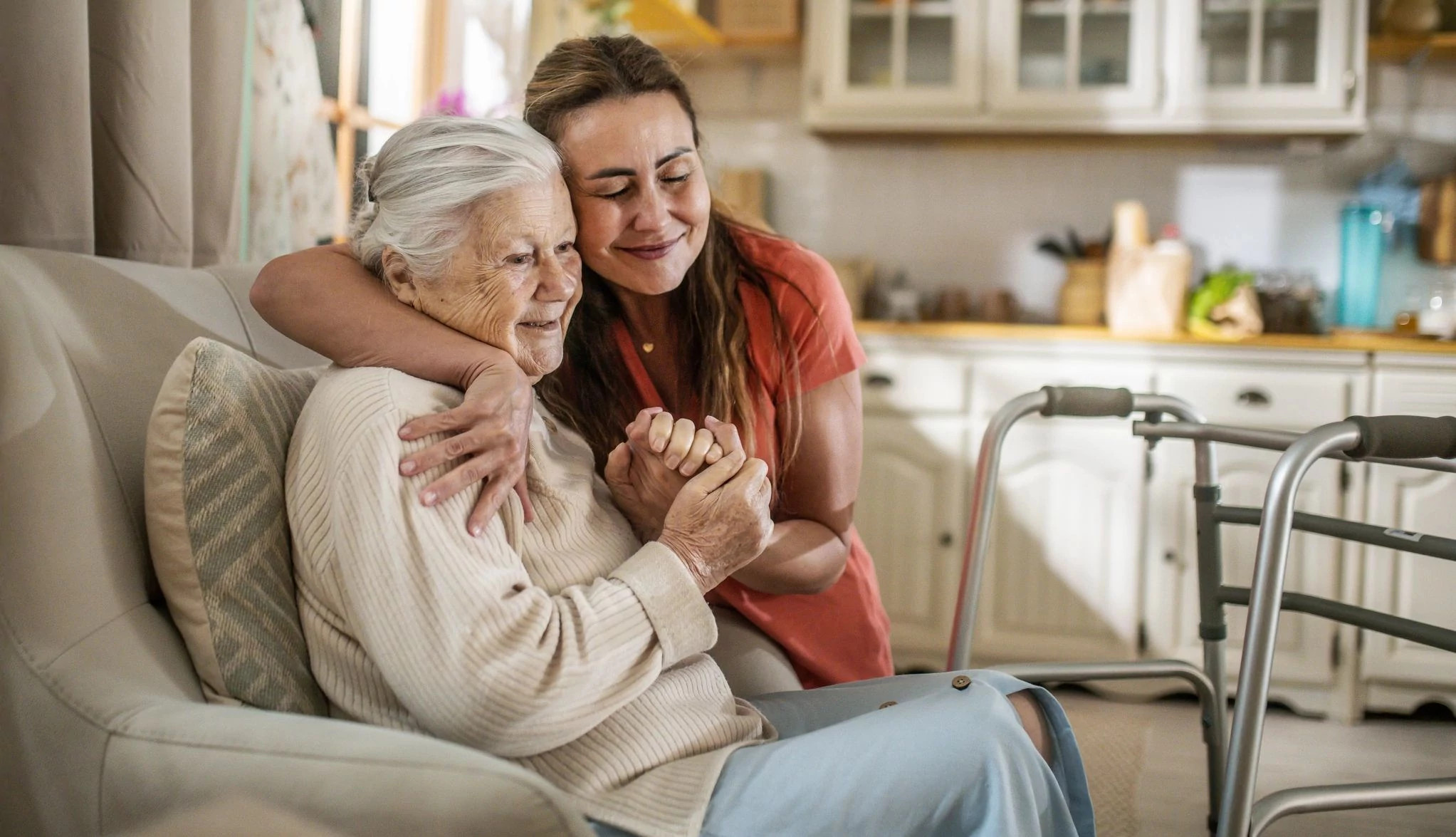 A woman smiles warmly while embracing an older female relative who is seated indoors, with a walker visible nearby in the room