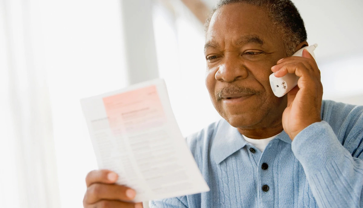 A man with a mustache talking on the phone about the bill in his hand.