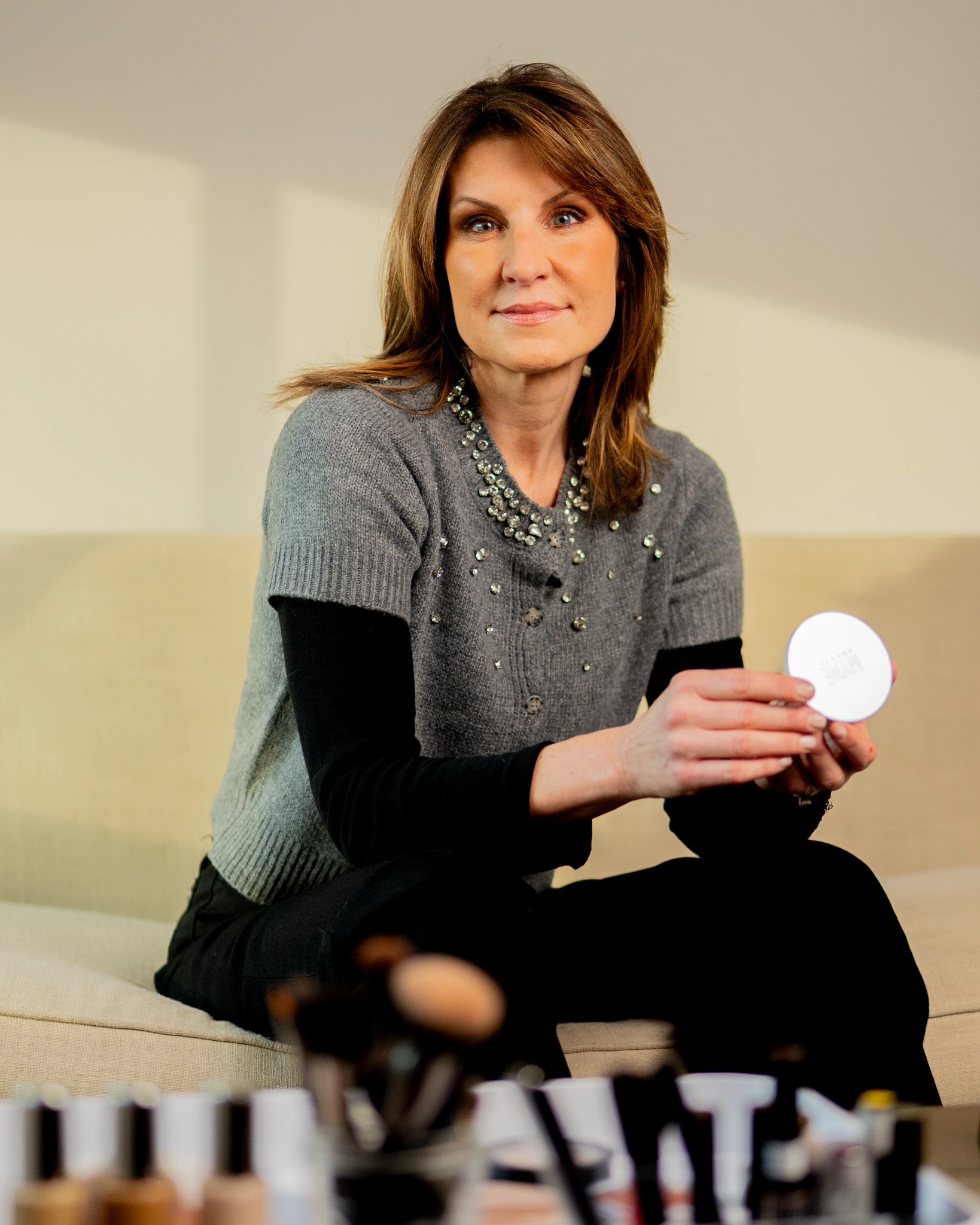 a woman poses while sitting on a couch and holding a compact