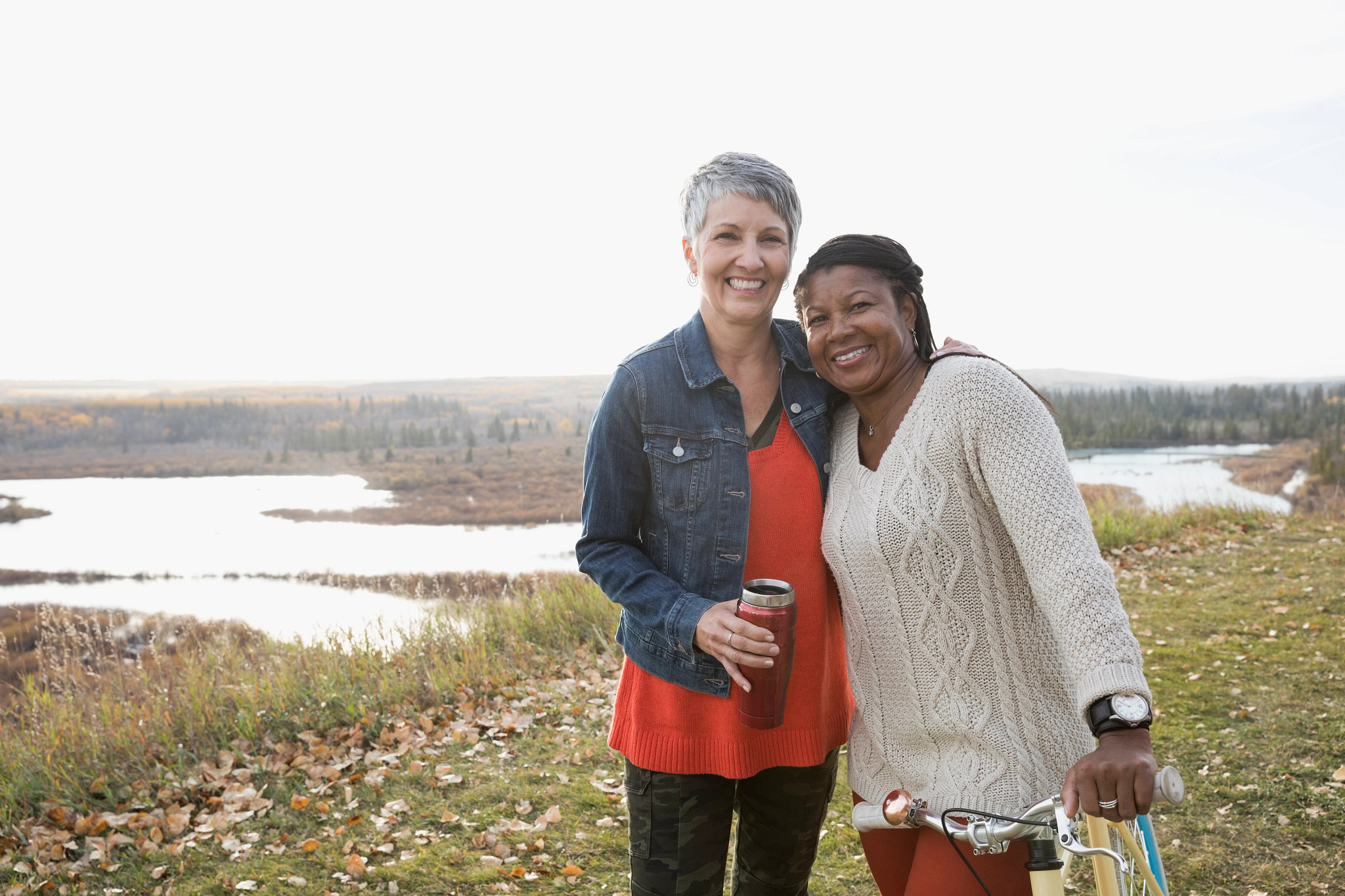 Smiling women with insulated drink container and bicycle on autumn hilltop overlooking lake
